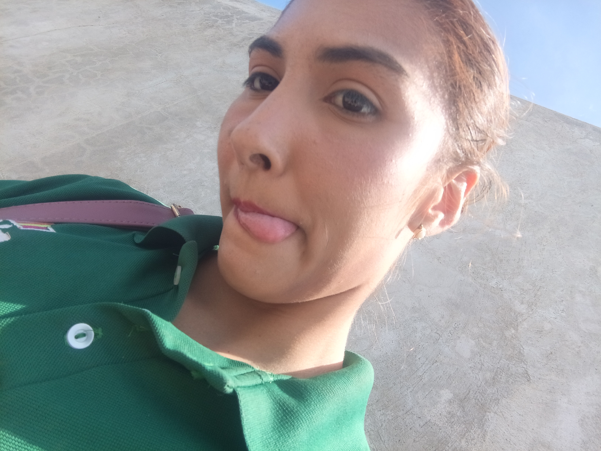 A medium-shot selfie depicts a young woman with light brown skin, dark eyebrows, and brown hair pulled back into a loose bun. She has light eye makeup and her tongue is sticking out in a playful manner. She is wearing a green polo shirt with white buttons. A pink strap is visible on her shoulder, suggesting a bag or purse. The background is a light gray wall, possibly made of concrete, and a sliver of bright blue sky is visible in the upper right corner. Considering the prompt's specification of Juan Griego, Venezuela, the photo likely depicts the woman in an outdoor location within that city, possibly near a building or structure with the gray wall. The light suggests a sunny day.