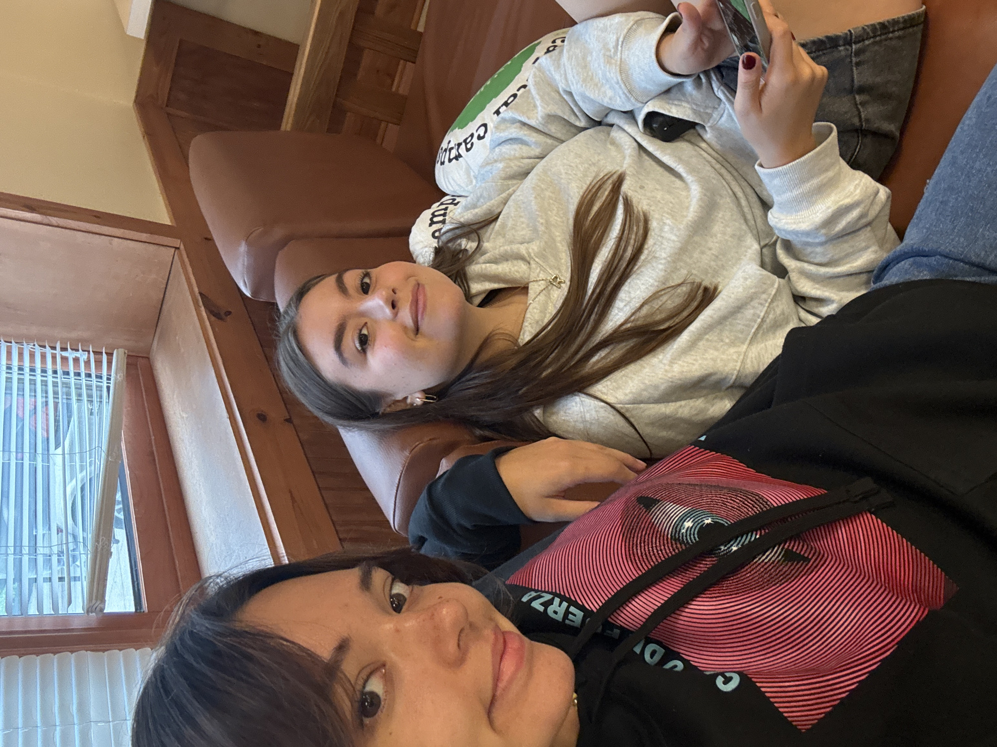 Two young women are sitting on a brown leather couch in what appears to be a room with wooden paneling and a window with blinds. The woman on the left is closer to the camera and is wearing a black hoodie with a pink and blue graphic design of an eye. She has dark hair and is looking at the camera. The woman on the right is wearing a light grey sweatshirt and is looking at her phone. She has long brown hair and is wearing a necklace. Both women appear to be relaxed and are likely at home or a similar casual setting. The location is identified as Washington Township, United States of America.