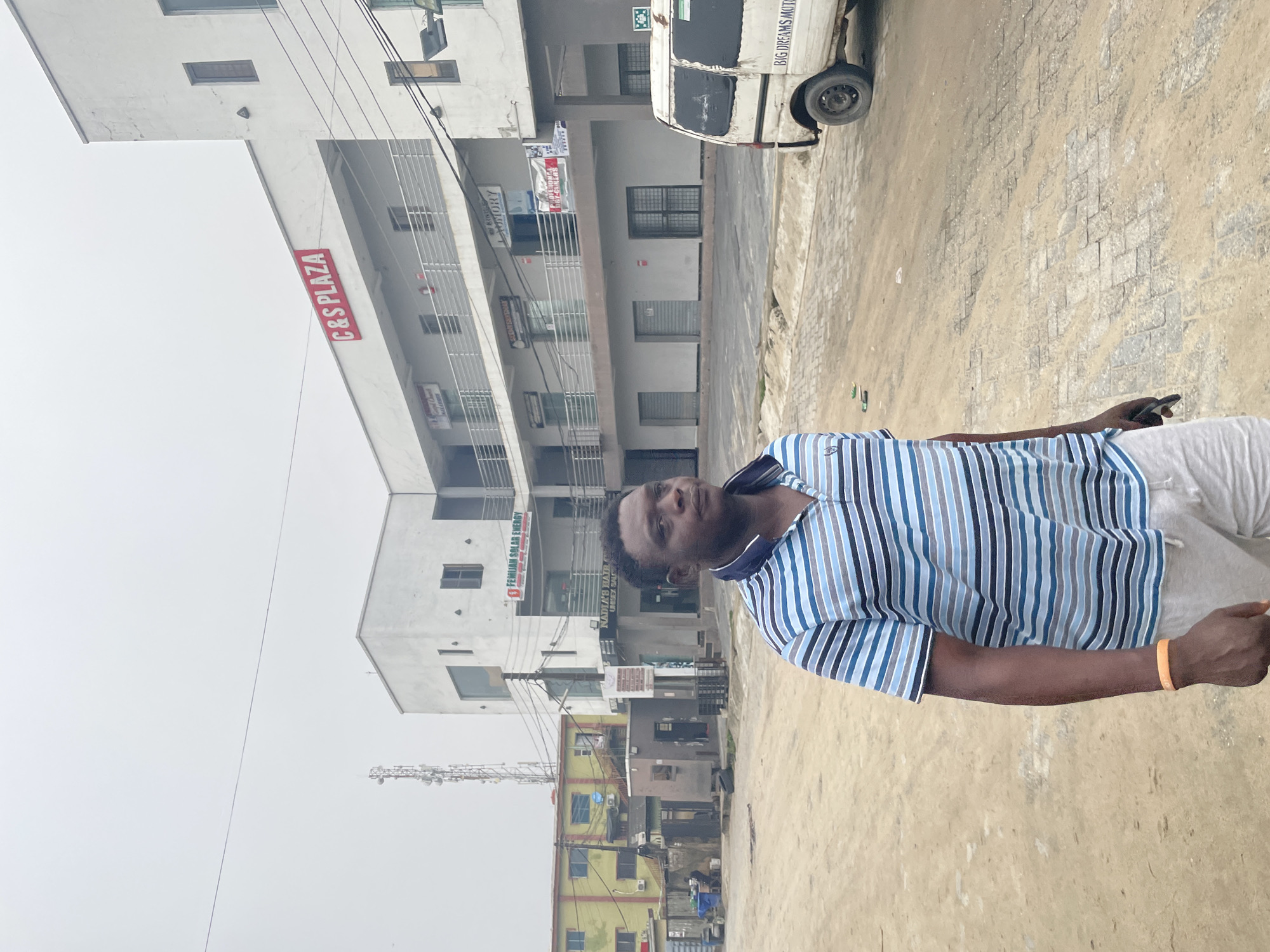 The image depicts an urban street scene in what appears to be a commercial or mixed-use district, consistent with areas found in Lagos, Nigeria.
In the foreground, an adult male of African descent is positioned slightly to the left of the frame's center, facing partially towards the right. He has a dark complexion, short dark hair, and is wearing a blue, white, and black horizontally striped polo shirt along with light grey shorts. An orange wristband is visible on his left wrist. He holds a dark object, possibly a mobile phone, loosely in his right hand. His expression is neutral, and his gaze is directed off-frame to the viewer's left.
The ground beneath him and extending into the midground is a mix of reddish-brown dirt and grey interlocking paving stones, indicating a partially unpaved or unevenly paved road surface. Scattered small pieces of green litter are visible on the ground in the midground.
In the background, a prominent white, three-story building, identified by a large red sign as "C&S PLAZA," dominates the right side of the frame. This building features multiple balconies with metal railings across its upper two floors. Various smaller commercial signs are affixed to its facade, including "FEMIJAN SOLAR ENERGY" and "NAIRA'S HAIR & BEAUTY SALON" on the lower levels, and "KING'S LAUNDRY PROFESSIONAL DRY-CLEANERS" on an upper level. Several ground-floor units feature rolled-down metal shutters or windows secured with metal grates.
To the left of the C&S Plaza, further in the background, stands an older, two-story building painted in yellow with red trim, which appears to be residential or mixed-use, displaying air conditioning units and laundry lines. Behind this yellow building, a tall, slender cellular or transmission tower is visible against the sky. Numerous black electrical or communication wires traverse the upper portion of the frame.
On the far right, a white commercial van, possibly a mini-bus, is parked with its rear facing the viewer. The words "BIG DREAMS MOTORS" are faintly visible on its rear. The sky above is overcast and uniformly light grey.