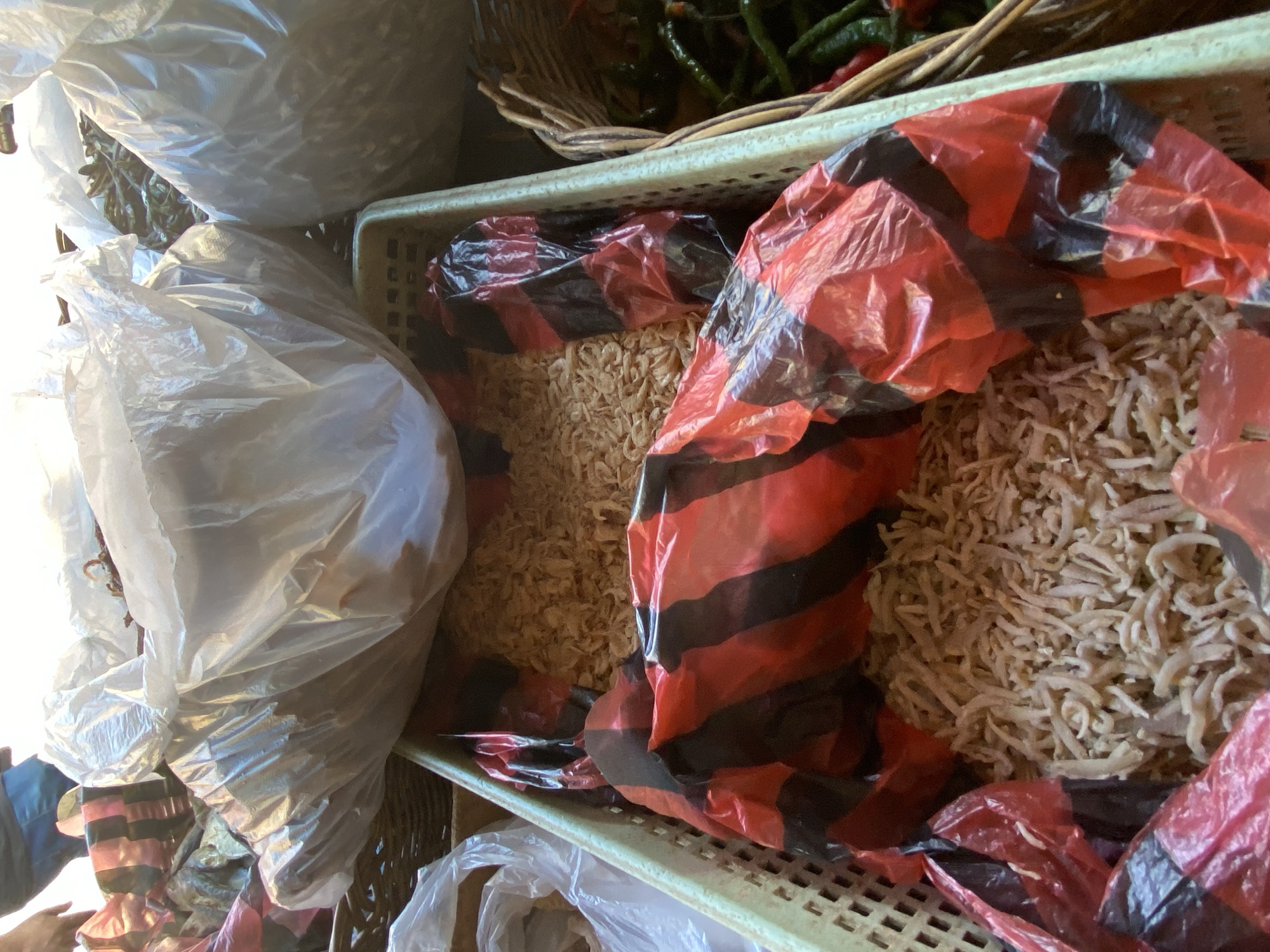 In an outdoor market setting, likely at a food stall, an assortment of goods is displayed. The foreground features a light-colored plastic crate filled with dried, small, light-brown shrimplike creatures. A red and black striped plastic bag is partially covering the contents of the crate, with some of the dried items peeking out from underneath. Behind this crate, larger white plastic bags, some appearing to be filled with dried goods, are piled high. To the right of the crate, a wicker basket holds a collection of green and red chili peppers. A person's legs, wearing blue shorts, are visible in the upper left corner, suggesting their presence at the market. The lighting suggests it is daytime.