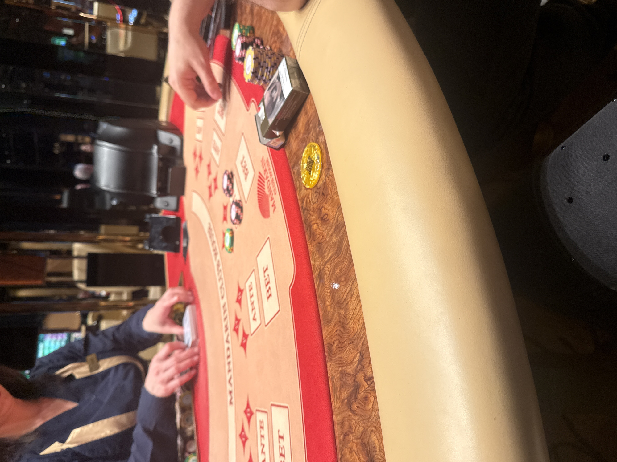 The image depicts a partially visible table at the Mandarin Club Casino in Batumi, Georgia. The table surface is covered with a red and beige cloth, imprinted with "MANDARIN CLUB CASINO" along one edge. It has designated spots labeled "ANTE" and "BET" for placing wagers. Several stacks of casino chips are visible, along with a deck of cigarettes in the upper-right of the frame. The chips consist of green, red, and gold chips, stacked on one another. A single gold chip is lying on the edge of the table. Two individuals are partially visible on either side of the table; one person is on the upper side and another person is on the lower side. The upper person is using their hands to move the chips. The lower person is holding cards and has a dark long-sleeved shirt. The table is bordered with brown wood on the outer area.