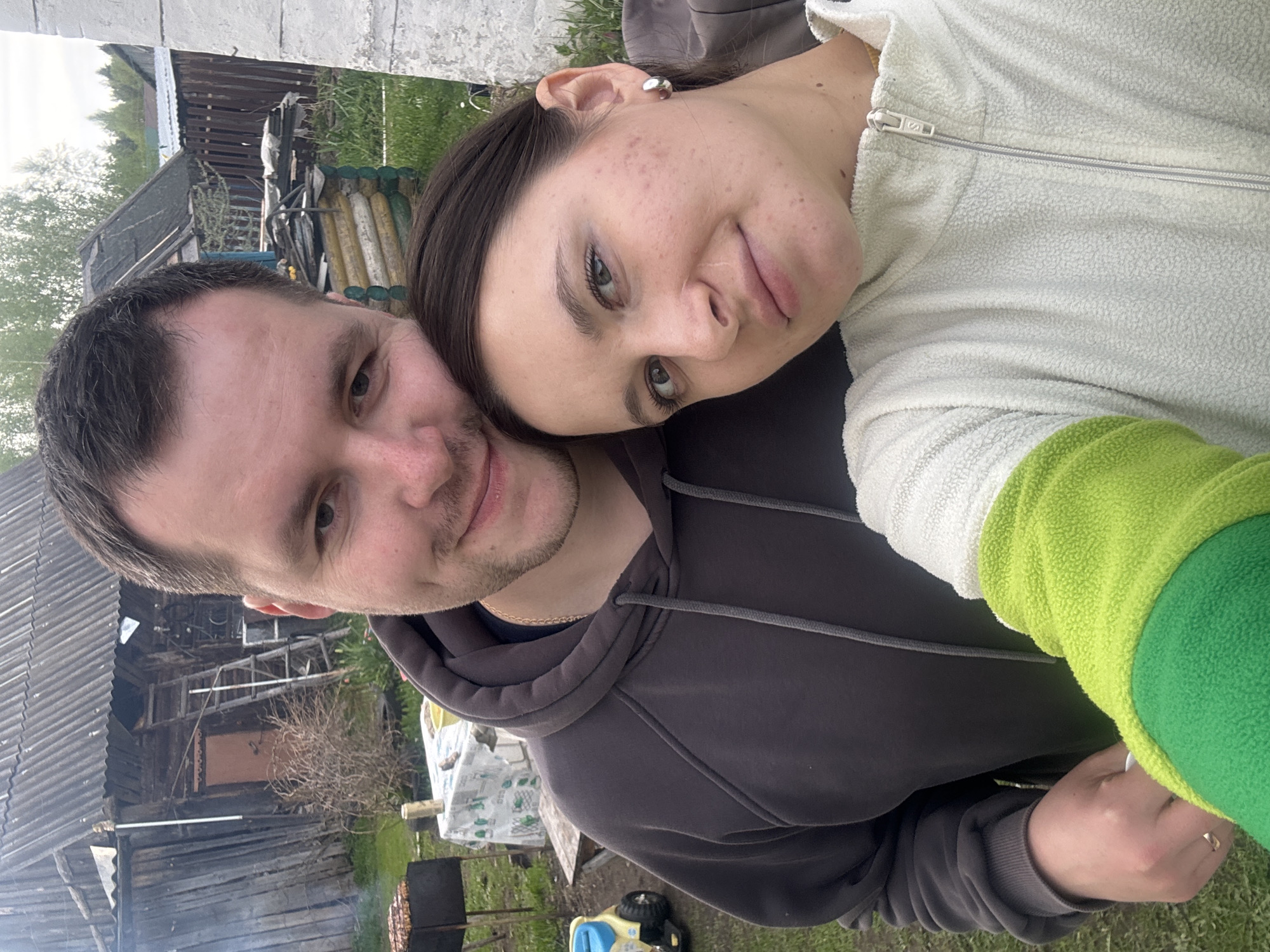 A selfie photograph shows a man and a woman posing closely together in an outdoor setting. The man, on the left, wears a gray hooded sweatshirt and has dark hair. The woman, on the right, wears a white fleece jacket with green sleeves. In the background, there is a rural landscape including a wooden house with a ladder visible, trees, and some stacked objects. There is also a grill visible in the grass. The photograph was taken in Красный Маяк, Russia.