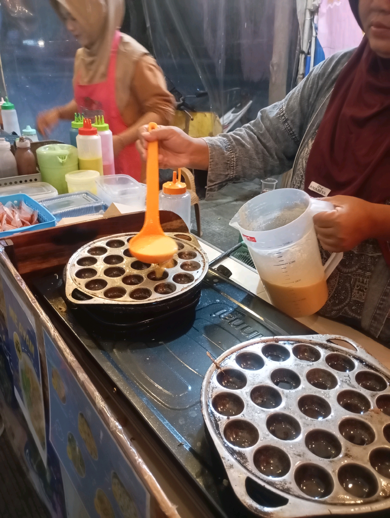 A close-up shot focuses on a street food vendor in Babelan, Indonesia, preparing a snack. In the foreground, a person's hands are visible, one holding a large plastic measuring jug filled with a light brown batter, and the other holding an orange ladle. The ladle is positioned over a circular cooking pan with multiple indentations, and batter is being poured into one of the indentations. The pan itself sits on a black stove. In the background, another vendor, wearing a beige hijab and a pink apron, can be seen working. Various bottles and containers of ingredients are lined up behind the cooking area, and a blue basket with pre-packaged food items is also visible. The overall environment suggests a bustling street food stall.