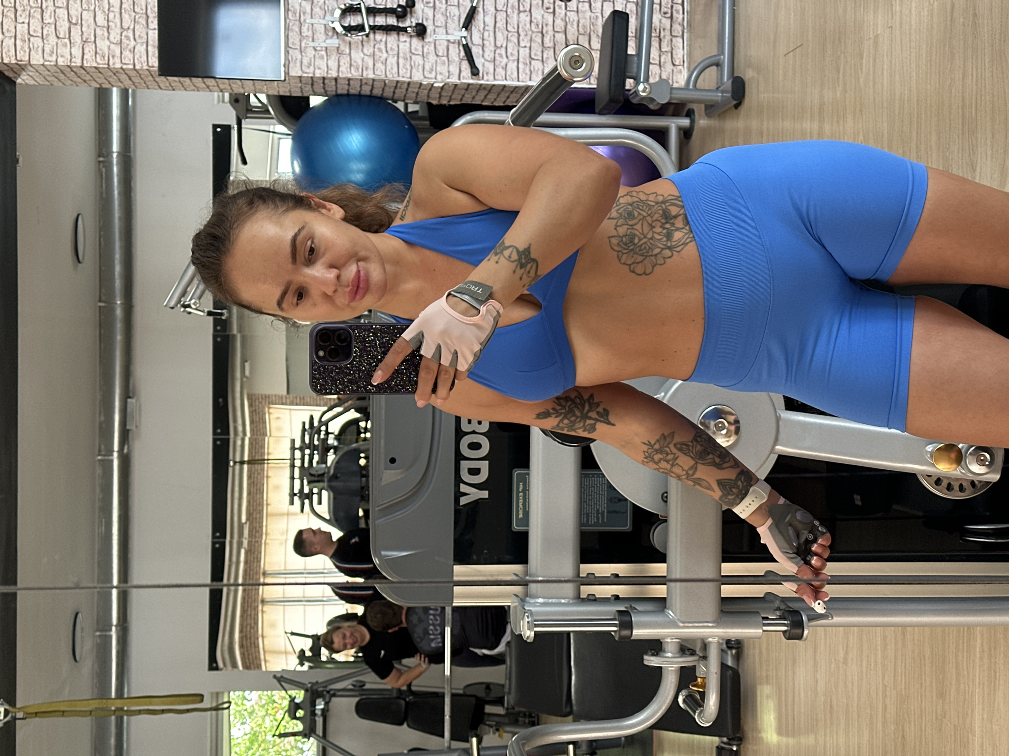 A woman with brown hair tied back in a ponytail is taking a selfie in a gym. She is wearing a blue sports bra and blue athletic shorts. She has tattoos on her arms and stomach. She is holding a phone in her right hand and an earbud in her left hand. In the background, there are gym equipment and mirrors reflecting other people. The gym appears to be modern and well-equipped. The floor is made of wood. The walls are painted in neutral colors and some have exposed brick. This scene takes place in the city of Yuzhnouralsk, Russia.