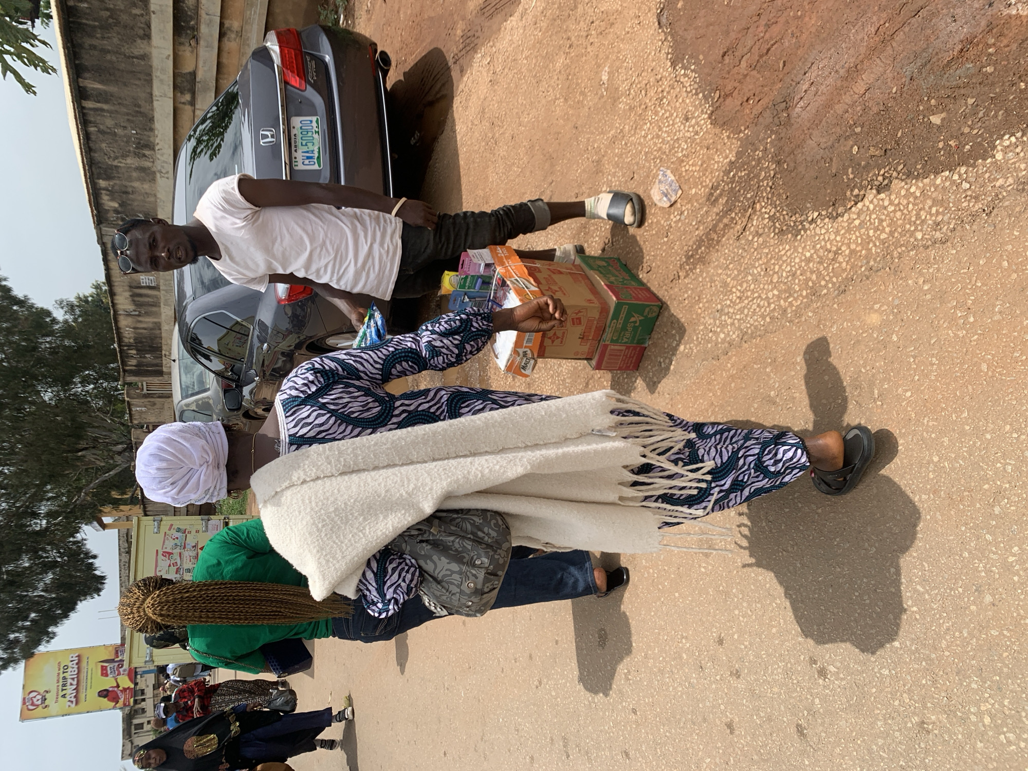 In Jos, Nigeria, an outdoor street scene features a young man standing by a car, tending to his small street vending stall which is piled with boxes and packets of goods, likely toiletries or small consumables. He is wearing a white t-shirt, dark jeans, and sandals. Behind him, a dark grey Honda Accord car is parked, its license plate clearly visible as "GWA 509DQ". The car is on a dusty, sun-drenched road with a rough, uneven surface. Several people are walking in the background, including two women, one wearing a green shirt and the other a colorful patterned dress, and a woman in a black hijab. In the distance, a large yellow billboard with red lettering advertises a trip to Zanzibar. The environment suggests a typical day in a busy, commercial area of the city.
