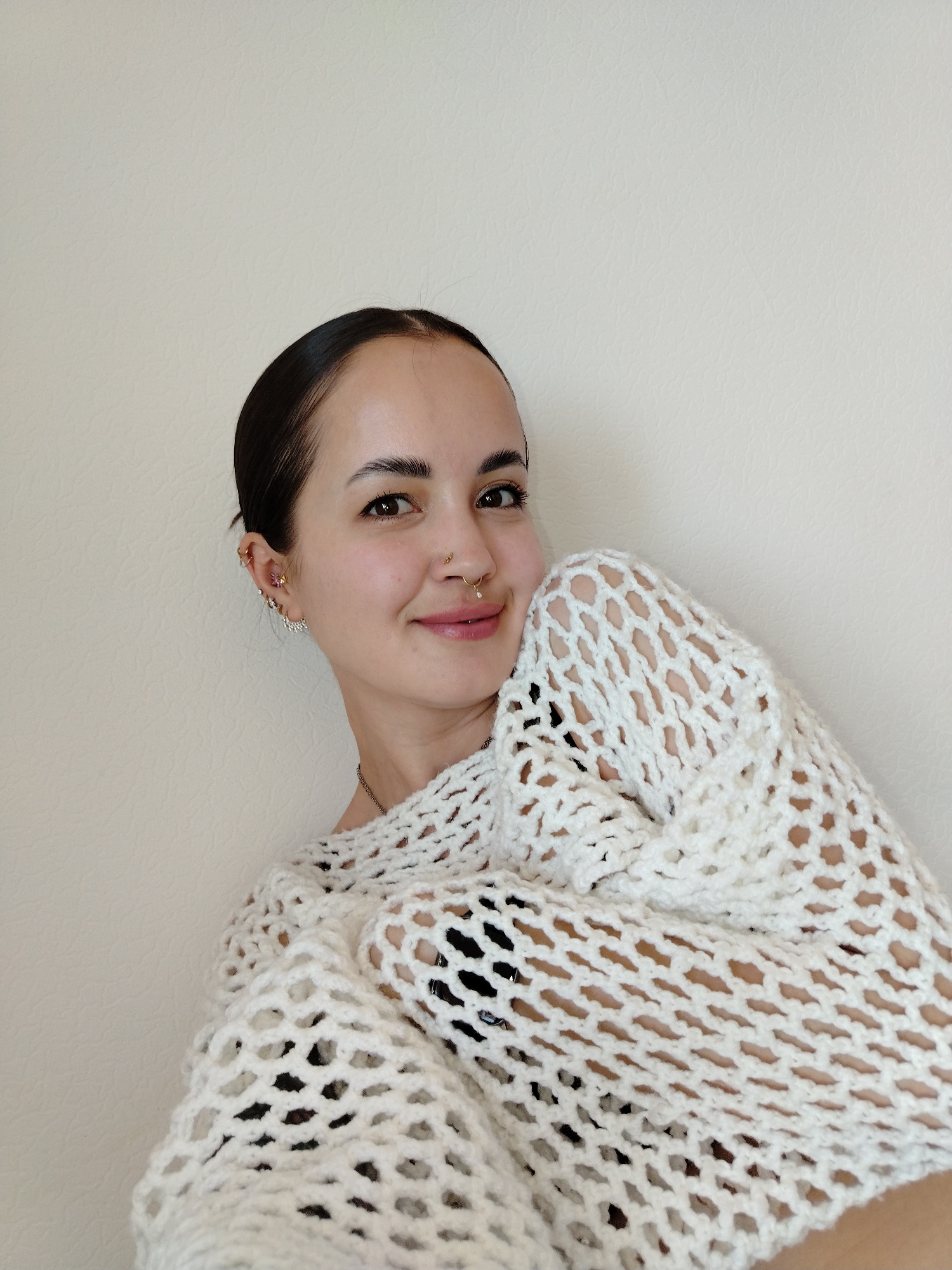 A young woman with dark hair pulled back in a bun is smiling at the camera. She has multiple piercings in her left ear and a nose ring. She is wearing a white, open-weave sweater. The background is a plain, light-colored wall. There is no indication of the location being in the city of Noviy, Russia.