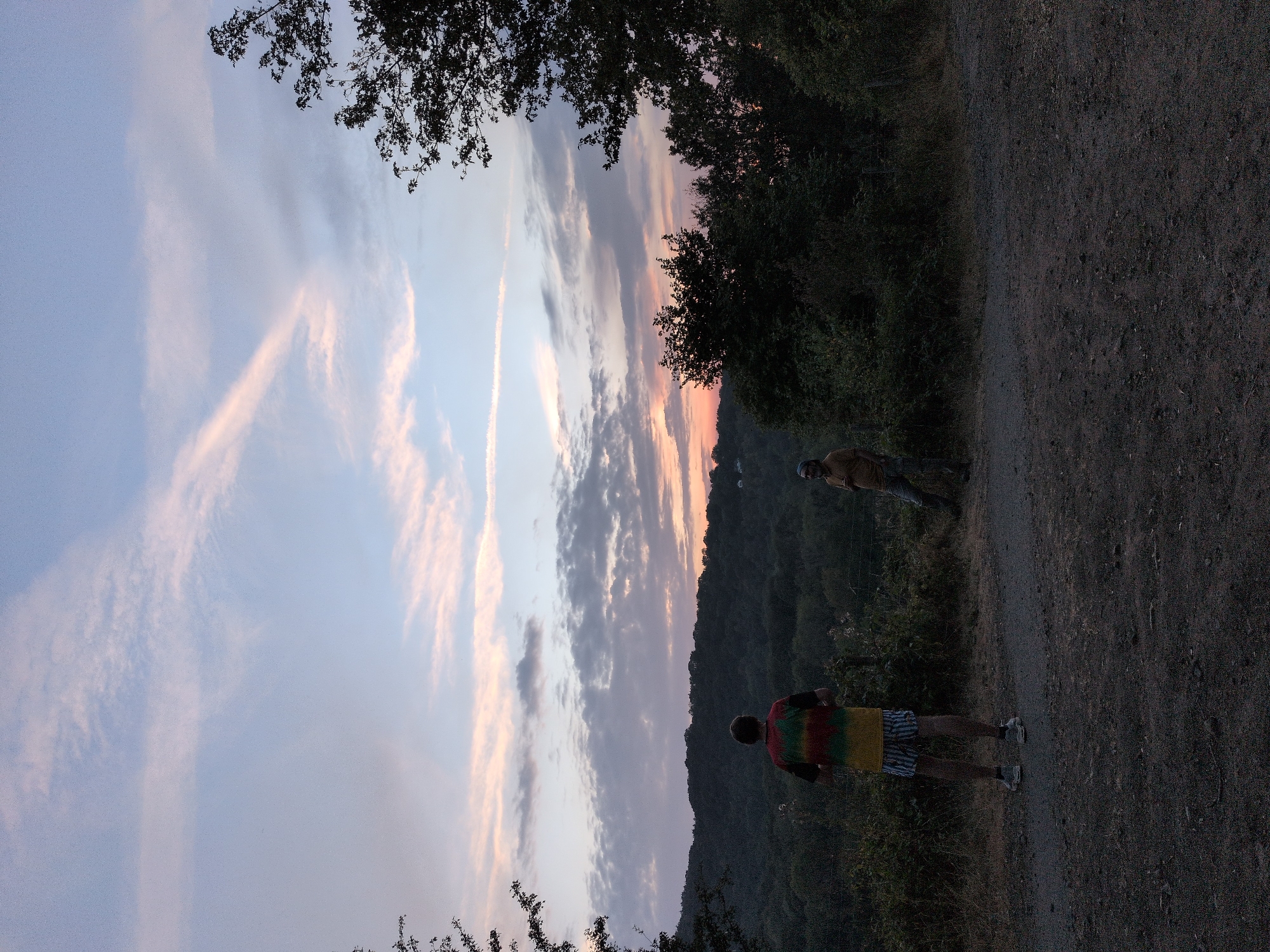 Two individuals stand in an open area facing a treeline, with a valley and a hill in the background. The sky is partly cloudy with hues of pink and blue, indicating either sunrise or sunset. The person on the left is wearing a tie-dye shirt and striped shorts, while the person on the right is wearing a tan shirt and camouflage pants. The ground is covered in gravel and dry grass. The location is Little Wenlock, United Kingdom.