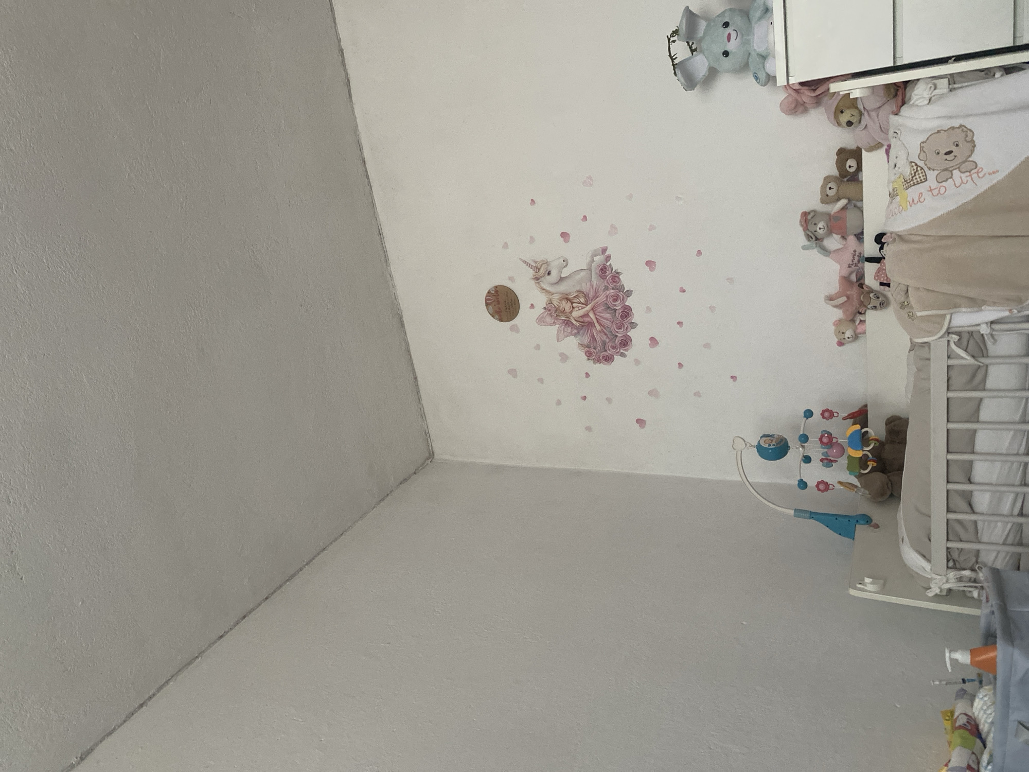 The image shows a room in a private dwelling. The walls are painted white and gray. On the ceiling, there is a unicorn decal surrounded by scattered heart decals. A baby crib is positioned in the right side of the image, and above it, there are stuffed animals, a baby mobile, and baby supplies. The location is likely in Bulgaria, based on the city name, but further confirmation would require more visual clues.