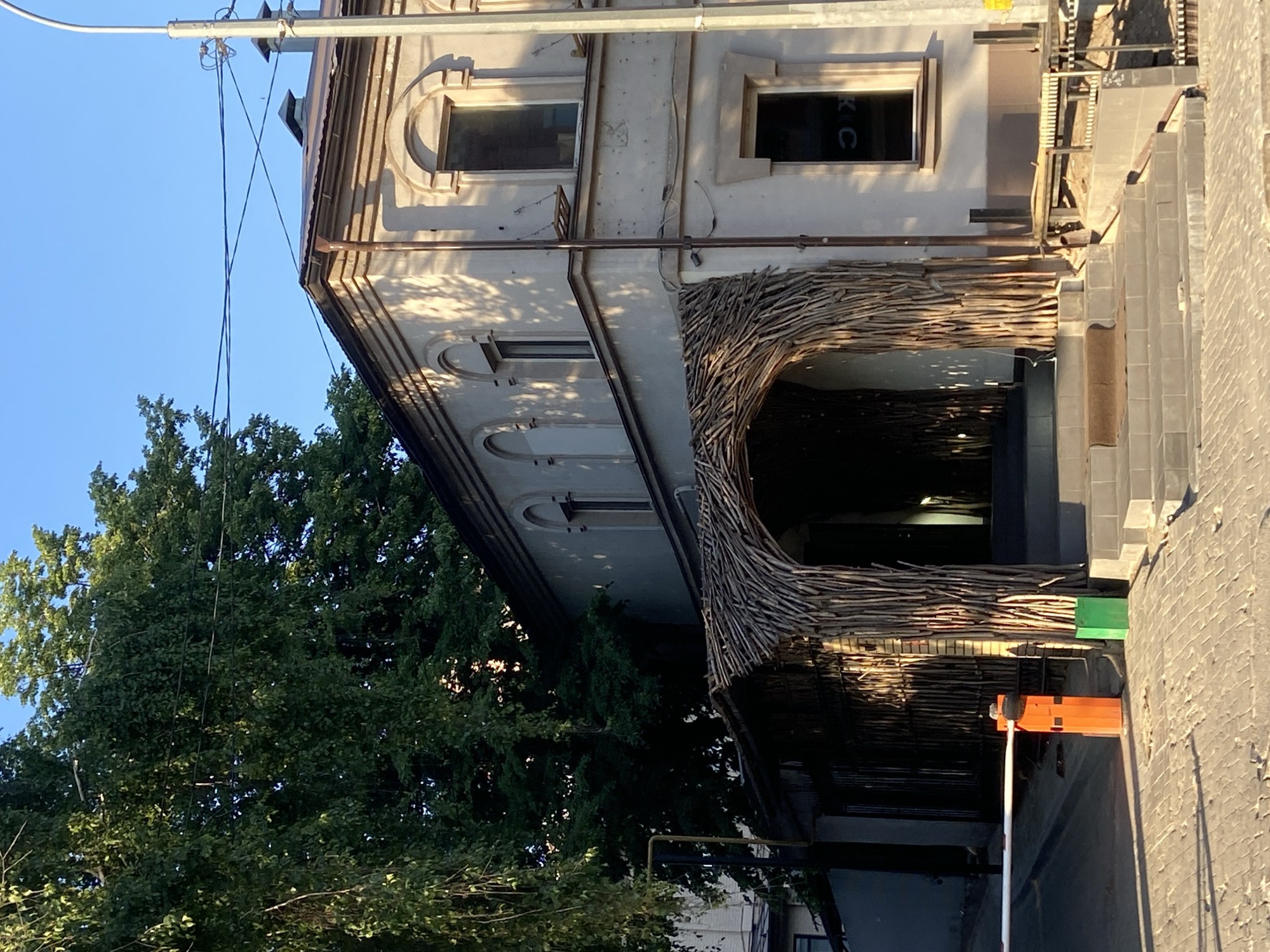 The image shows a building with a unique entrance in Kazan, Russia. The entrance is adorned with an arch made of interwoven branches, creating a nest-like appearance. The building itself has a light-colored facade with arched windows and classical architectural details. To the left of the building, a large tree with dense foliage provides shade. A utility pole with wires extends into the clear blue sky above the building. The sidewalk in front of the building is cracked and uneven.