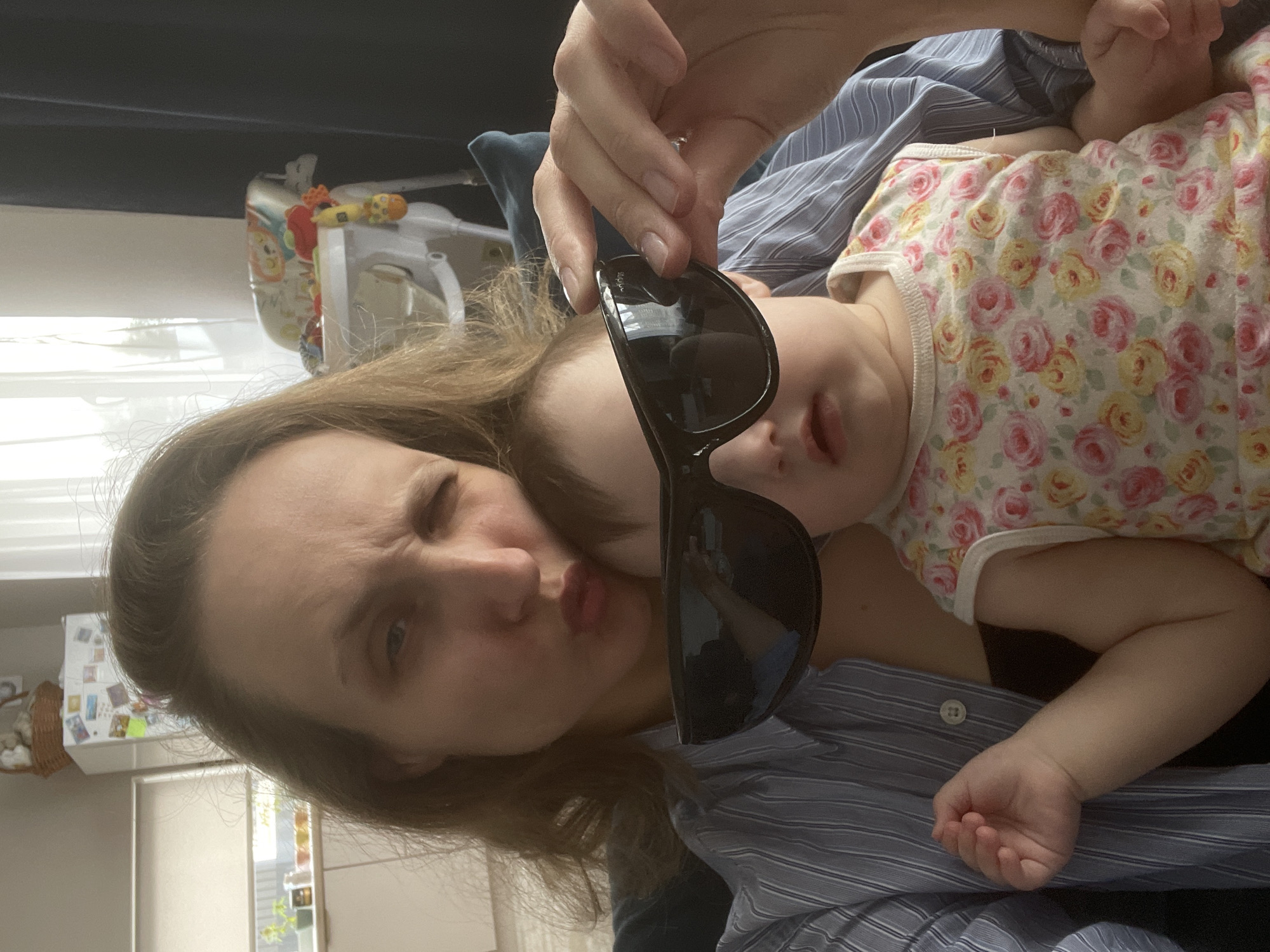 The image shows a woman holding a baby. The woman is looking at the camera with one eye closed and her lips pursed, as if posing for a selfie. In her other hand, she holds a pair of black sunglasses near the baby's face. The baby is lying on what appears to be the woman's chest, wearing a floral print onesie. The woman is wearing a blue and white striped shirt.
In the background, there is a baby's high chair, a white cabinet with various items on it, and a window. Based on the environment, it appears to be an interior setting, possibly a home. There is no indication of being in the specific city of Подшивалово, Russia.
