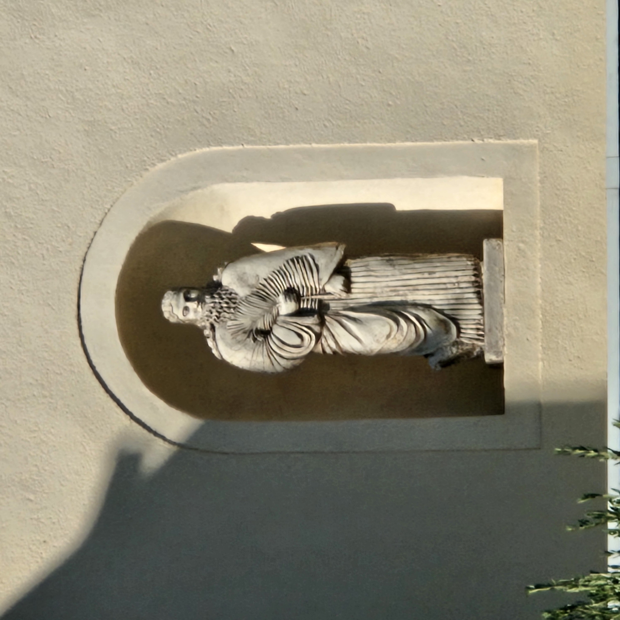 The image shows a stone statue of a man in classical attire, holding a fan, set within a recessed archway on a textured beige wall. The statue is depicted from the waist up, with detailed carvings of his beard, robes, and the fan. A shadow of the statue and archway falls on the wall to the left. In the lower foreground, blurred green foliage is visible. The statue is located on the facade of the Narbonne Cathedral in Narbonne, France.