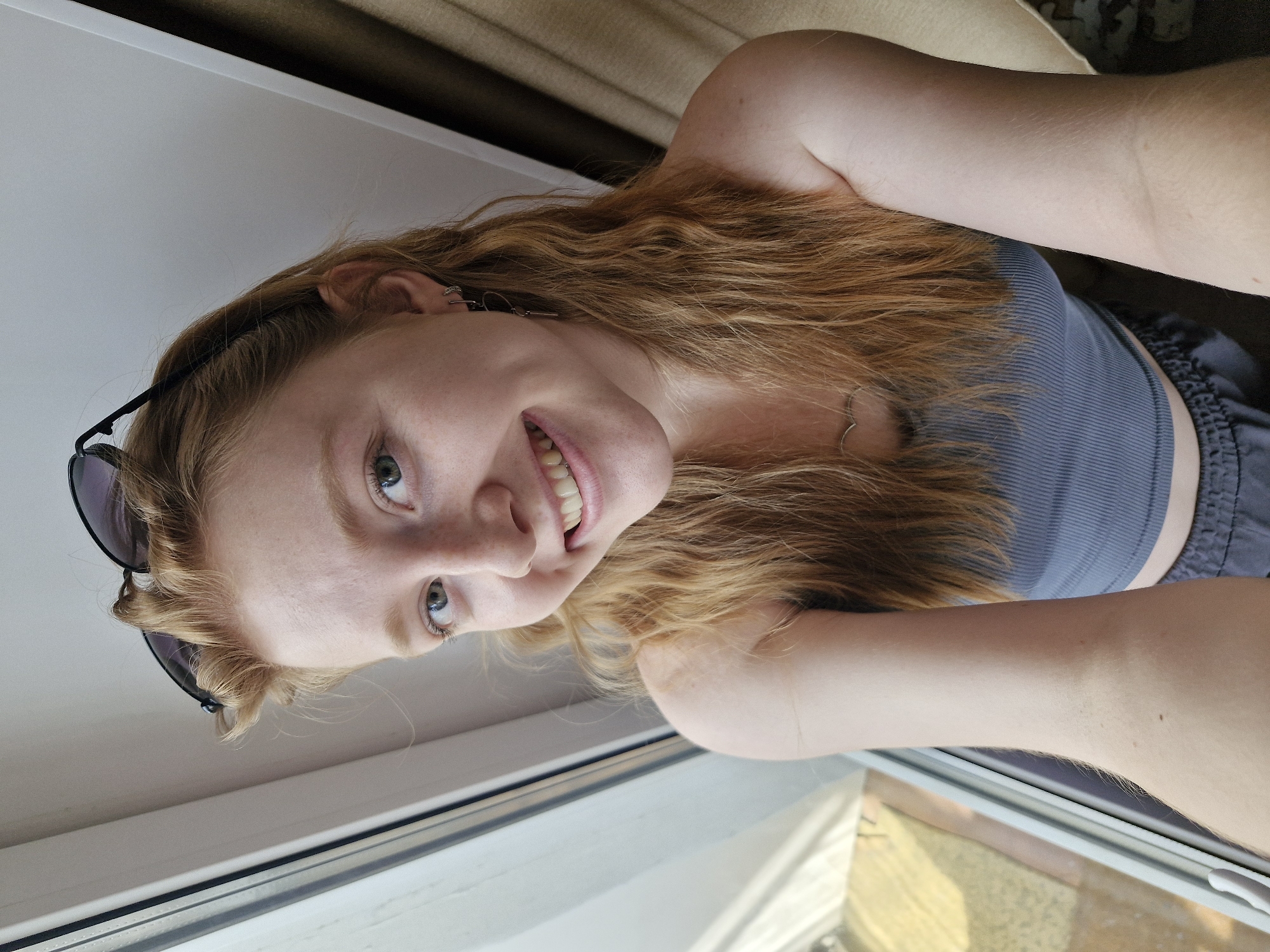A fair-skinned young woman with light brown/red hair is shown from a high angle in a selfie. She's wearing a gray top and dark shorts. Sunglasses are perched on her hair. She is smiling at the camera. A window is visible with sunlight coming through. There is no evidence of this photo being in Yekaterinburg, Russia.