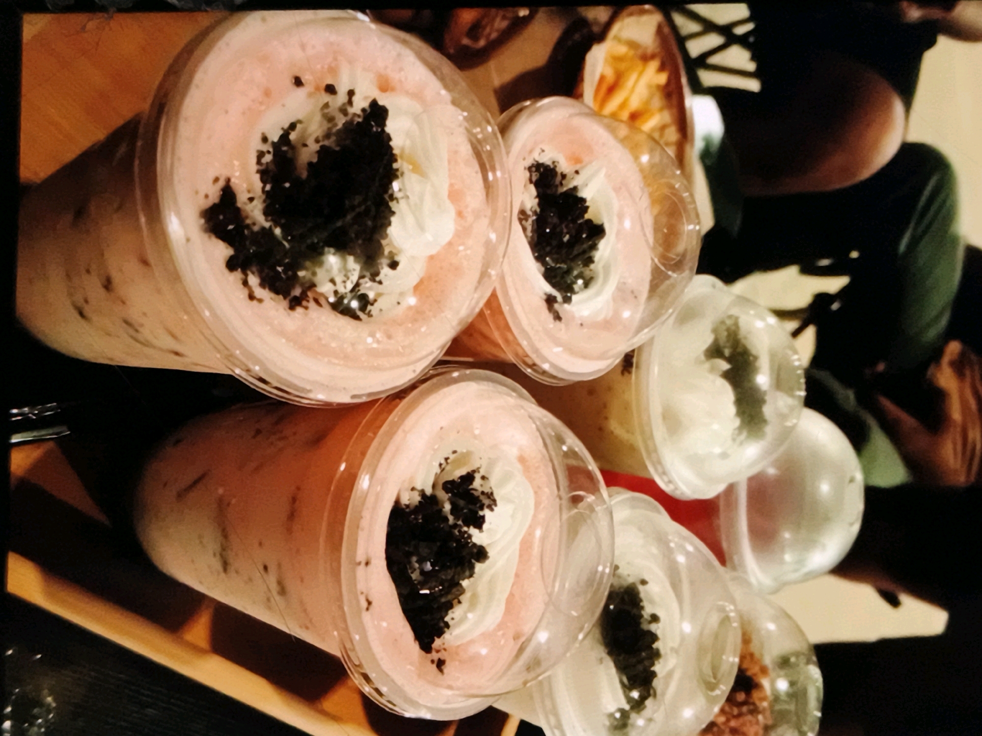A close-up, overhead view shows several clear plastic cups filled with pink beverages topped with whipped cream and dark crumbled toppings. The cups are arranged on a wooden surface. To the right of the cups, a bag of french fries is visible, along with parts of a person wearing a dark green shirt. The scene is dimly lit, suggesting an indoor setting. The location in Caloocan, Philippines, cannot be determined from the visual information provided.