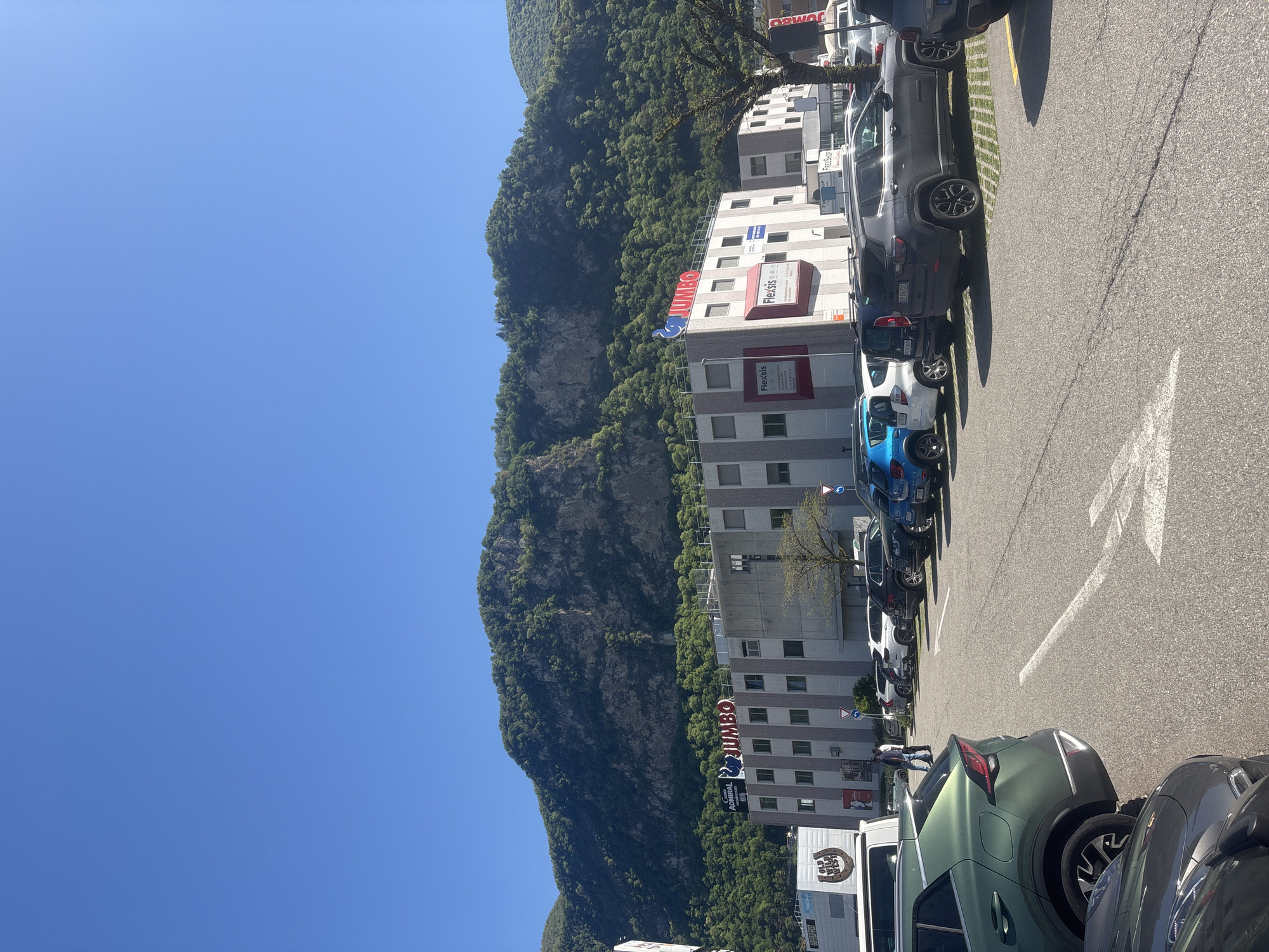 The image captures a bright, sunny day in Mendrisio, Switzerland, depicting a commercial or retail park area. In the foreground and midground, a parking lot is filled with numerous parked cars of various models and colors, including dark grey SUVs, a white hatchback, a blue car, and a green SUV. White parking lines and a directional arrow are visible on the asphalt.
Adjacent to the parking lot are several multi-story commercial buildings. A large, forested mountain forms the prominent backdrop, rising steeply behind the buildings. The sky is clear blue.
Visible text on the buildings includes multiple large red "JUMBO" signs. Other signs on the building facades show "Flexsis," "Admiral," "Pirellas," and possibly "Ferrovia." A white delivery truck or van parked in the lot has "GIAC. VISO" and "RAGUSA" printed on its side. At least one person is visible walking near the parked cars. The strong shadows cast by the vehicles and buildings suggest it is mid-day or early afternoon.