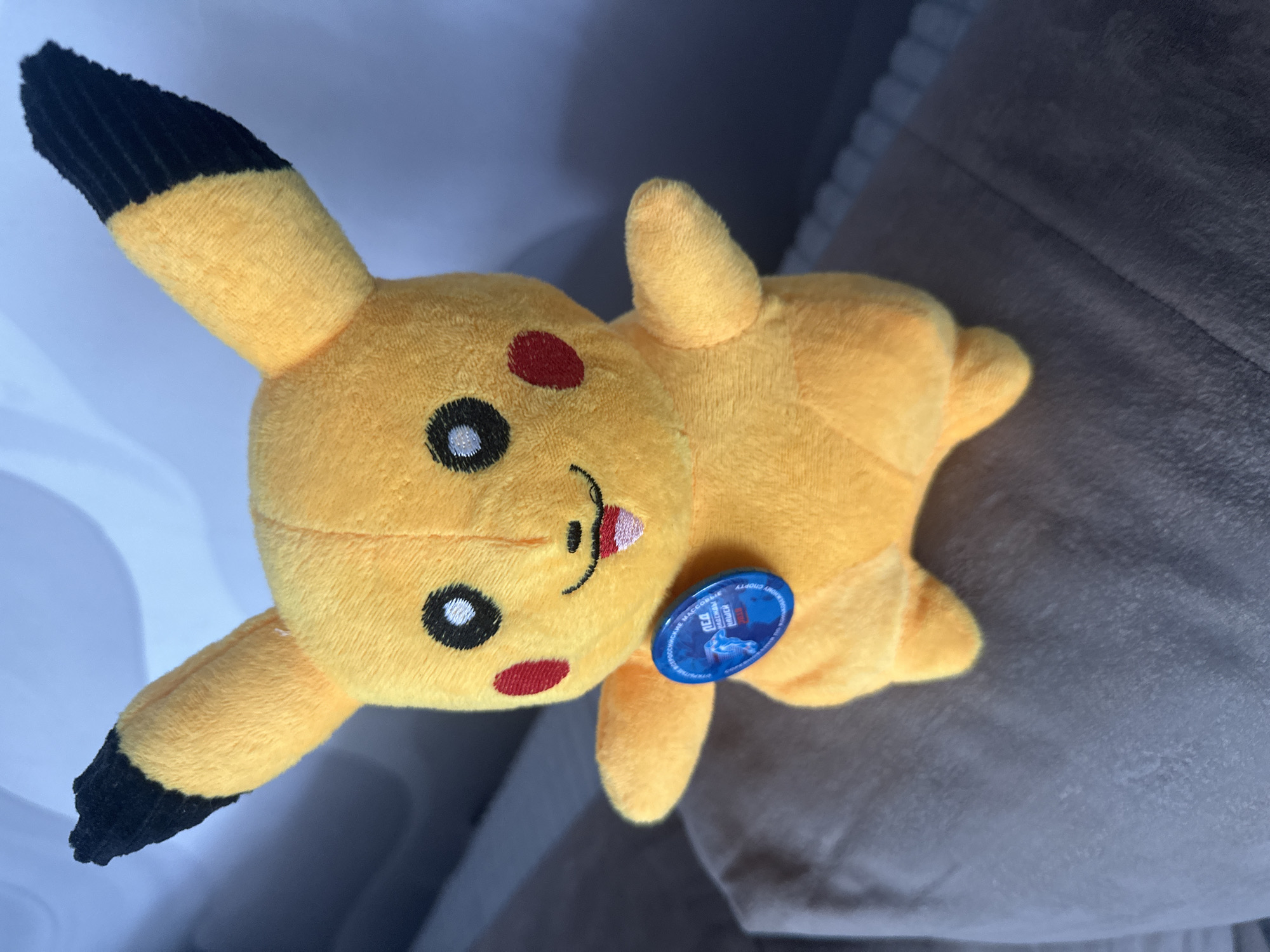 A plush toy Pikachu, the popular Pokémon character, is positioned on a textured grey surface, possibly a sofa or cushion, indoors. The toy is predominantly yellow, with black tips on its pointed ears, and features embroidered black eyes with white highlights, red circular cheeks, and a small, smiling mouth with a pink tongue. Attached to the toy's chest is a round, blue badge with text and an image that is not clearly discernible. The background is out of focus, showing a light-colored wall or surface, suggesting an indoor environment. There is no information in the image to indicate its location in Birobidzhan, Russia.