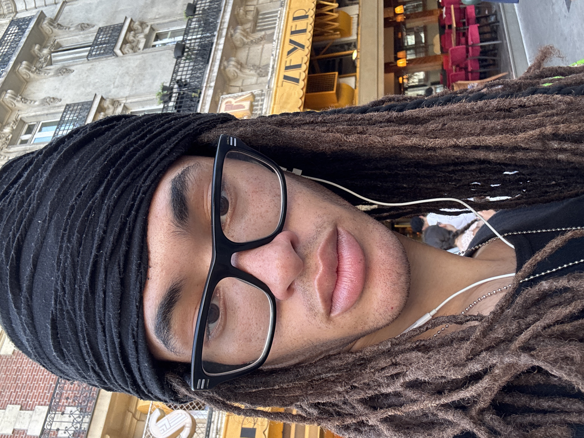 A man with dreadlocks and glasses is centered in the image. He is wearing a black head wrap and black-rimmed glasses. White headphones are visible, with the wire running down his neck. The background is out of focus, but it appears to be a street scene in Paris, France, with a building labeled "ZEYER" and a Parisian-style building with wrought iron balconies.