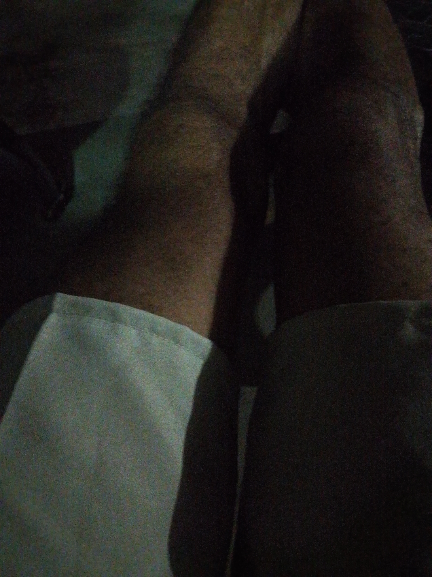 The image depicts a person's legs resting on a surface, possibly a bed. The person is wearing light-colored pants. The lighting is dim and creates a shadow effect. The location is not discernible from the image.