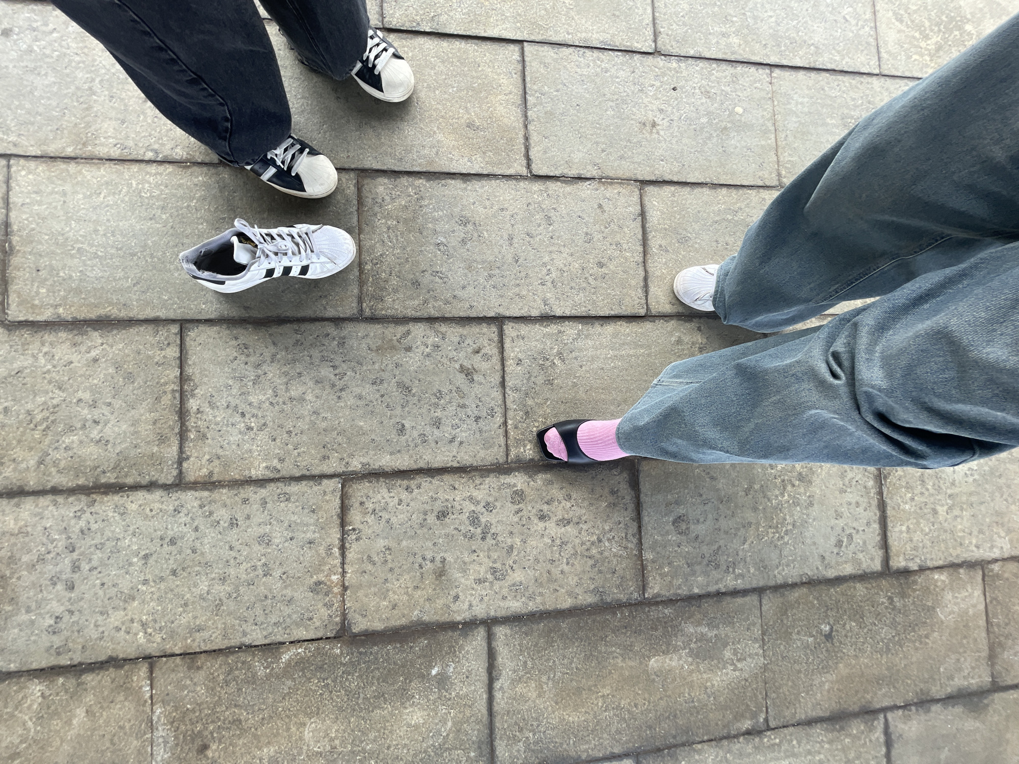 The image shows the lower legs and feet of at least three individuals standing on a paved ground made of large, rectangular stone tiles. The ground appears worn and slightly speckled.
Two people are visible in the upper left of the frame. One person is wearing dark wash jeans and black and white sneakers. The other person is wearing dark wash jeans and a pair of black and white sneakers. A single white sneaker with black stripes is placed on the ground to the left of the person wearing dark jeans.
In the lower right of the frame, another person is visible. They are wearing light blue, wide-leg jeans and white sneakers. Next to them, another person is wearing the same light blue jeans and black slides with pink socks.
The image seems to be taken from a high angle, looking down at the feet. The lighting suggests it could be daytime, with no strong shadows or indications of specific weather. The setting appears to be an outdoor public space, possibly a walkway or plaza. There is no visible text in the image. The overall impression is casual, perhaps a group of friends or companions gathering.