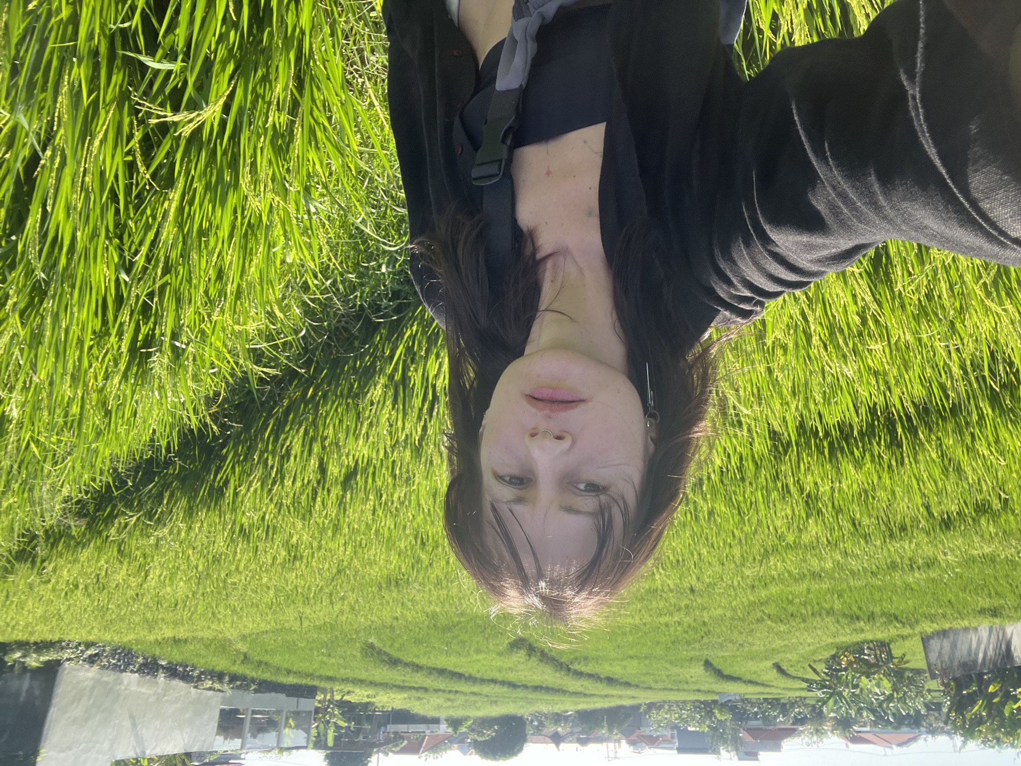 A woman with brown hair, wearing a black top and jacket, is situated in a lush, green rice field in North Kuta, Indonesia. The field stretches into the distance towards a residential area with buildings visible on the horizon. The photo appears to have been taken at ground level. The subject is facing toward the camera with the rice paddy filling the foreground and background.