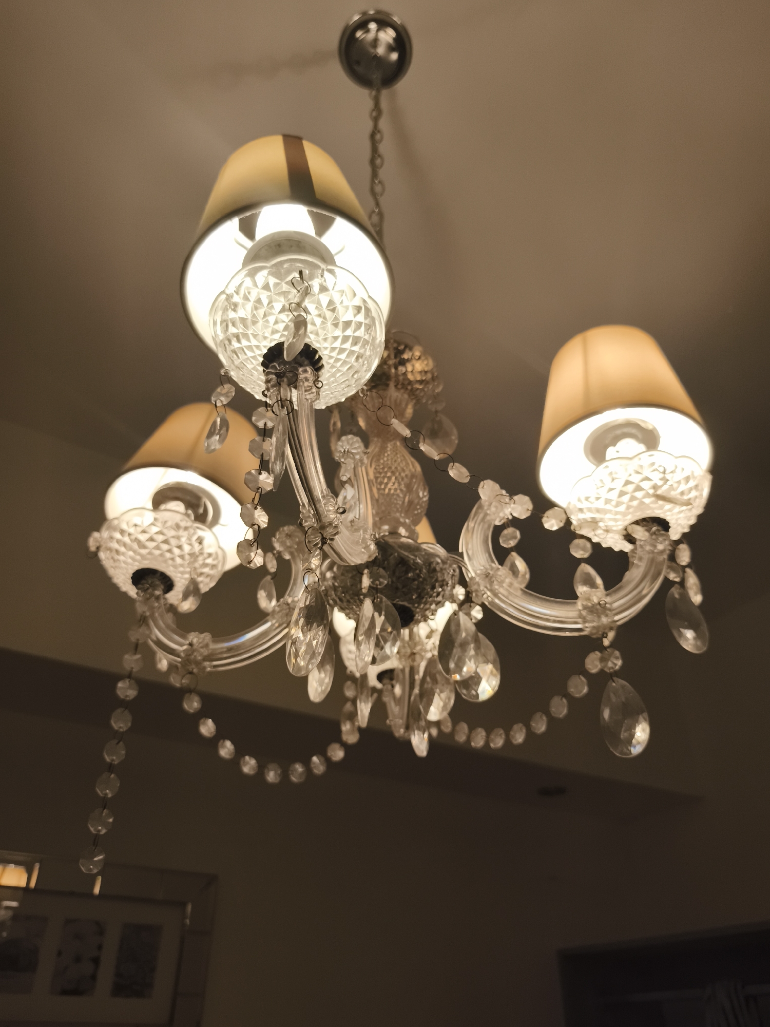 The image shows a chandelier with three lampshades and crystal accents. The lampshades are beige with a dark band at the top. The light is warm, suggesting a residential setting. The location is not discernible from the image.