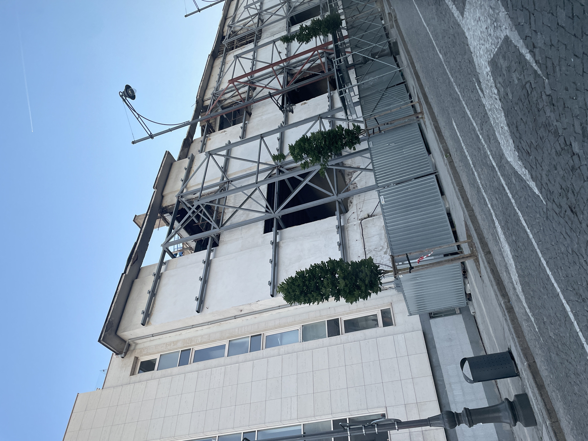 The image depicts a building under construction or renovation in Sofia, Bulgaria. The building has a multi-story facade, with a combination of newer tiled sections and older, damaged sections with exposed structural elements. Scaffolding supports the damaged sections. Patches of green vegetation grow from the scaffolding. A street with a cobbled surface is visible on the right side of the frame. A street lamp and litter bin stand next to the pavement. A blue sky is visible in the background.