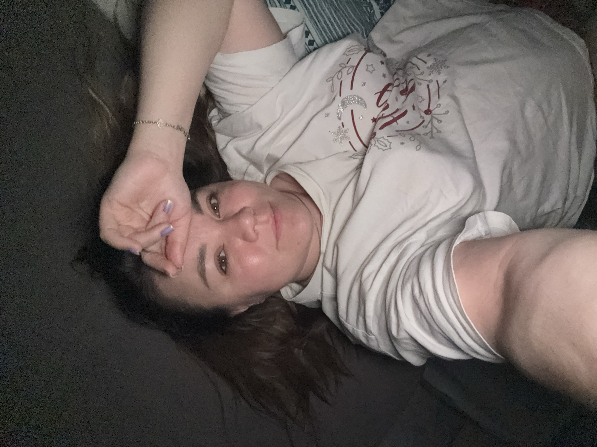 A woman with brown hair is lying on her side on a bed, with her head turned towards the camera. She has her right hand on her forehead and is wearing a white t-shirt with a holiday-themed design. The background is dark and blurry, suggesting a dimly lit room. The image appears to be a selfie, taken from a low angle. There is no information available about the location in the city of Stary Oskol, Russia.