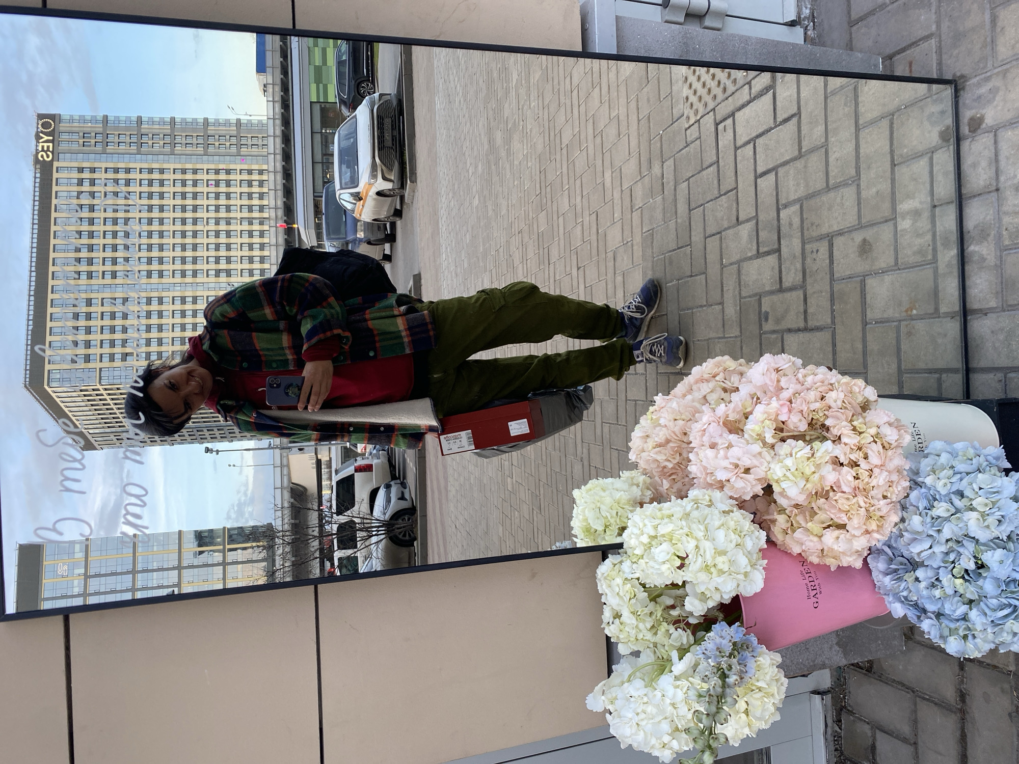 The image shows a reflection in a large, rectangular mirror. The reflection depicts a person holding a phone, wearing a plaid shirt, red t-shirt, and green pants. They are standing on a paved sidewalk. In the background of the reflection, there is a building labeled "ОЛЕГ" and cars on the street. To the right of the mirror, there is an arrangement of artificial flowers in pastel colors, including pink, white, and blue, placed in a pink bag labeled "Home Life GARDEN With You." The mirror appears to be leaning against a wall or surface, with a section of the wall visible below it. Based on the building label "ОЛЕГ," the location is Moscow, Russia.