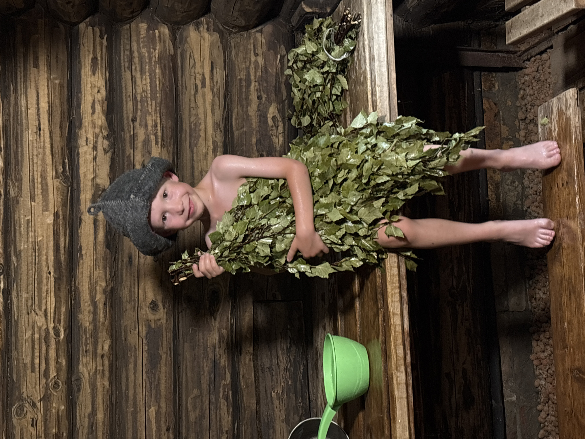 A young child wearing a grey sauna hat is lying down in a wooden sauna in Vanyki, Russia. The child is partially covered by a bundle of birch branches, commonly used in saunas. The child is looking towards the camera with a slight smile, appearing relaxed. One foot is visible, resting on a wooden bench. To the right, a green ladle is positioned over a bucket. The sauna walls are made of rough, dark wood, and there are stacks of stones in the background. The lighting suggests it could be daytime, but the interior of the sauna is dimly lit. The overall scene conveys a sense of tradition and relaxation associated with Russian sauna culture. There is no visible text in the image.