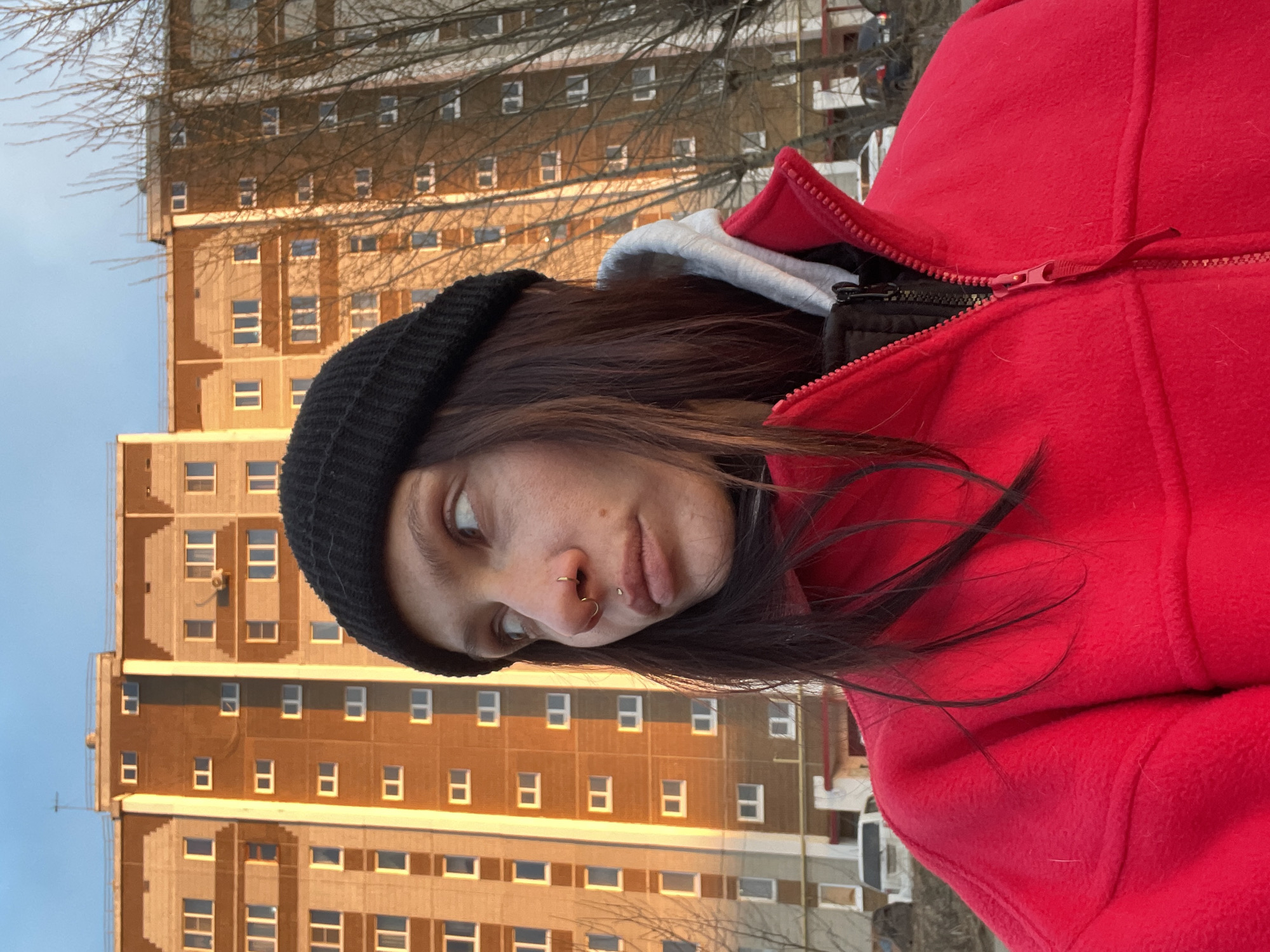 A person is depicted in a close-up, outdoor shot against an urban backdrop. The subject, on the right side of the frame, has dark hair and wears a black knit beanie, a bright red fleece jacket, and a grey hoodie underneath. They have a gold-colored septum ring and a silver-colored philtrum piercing. Their gaze is directed upwards and to the left, with a pensive or observant expression.
The background features a tall, multi-story residential building with a light brown and grey facade and numerous rectangular windows. The building is illuminated by a warm, golden light, suggesting either late afternoon or early morning. Bare tree branches are visible in the foreground, indicating a cold season. The sky is clear and light blue. The scene depicts an urban or suburban environment, consistent with Izhevsk, Russia. No visible text is present.