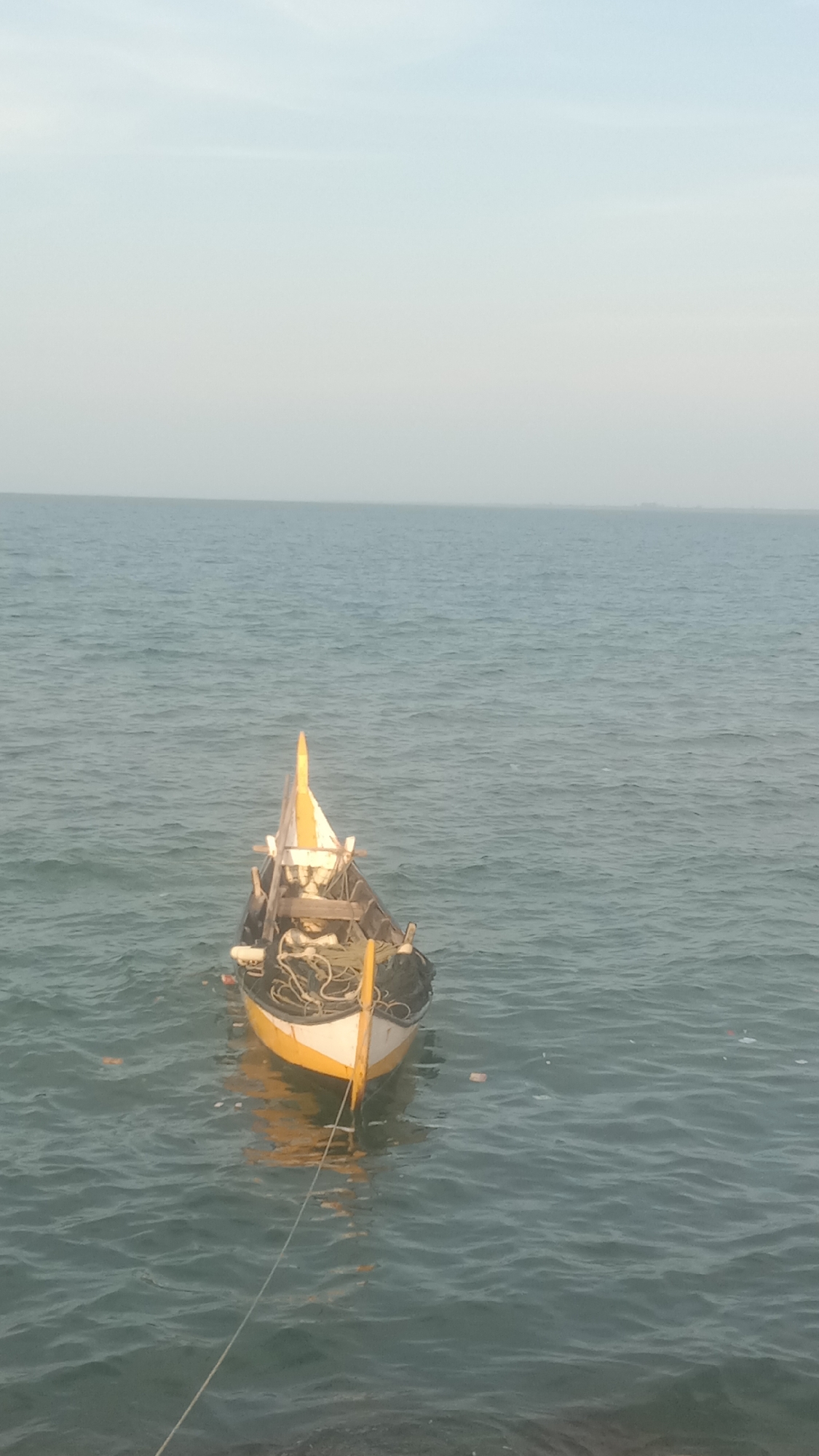 The image captures a small, traditional-looking fishing boat gently floating on calm, rippling sea waters under a hazy, light blue sky. The boat, positioned centrally, features a distinctive yellow and white painted hull at its bow, with a prominent upward-pointing yellow fin or decorative element at the very front. The rest of the boat appears to be dark-colored, likely wood, and is filled with fishing gear, including coils of rope and nets.
A rope extends from the boat's bow towards the bottom of the frame, presumably mooring it. Gentle reflections of the boat are visible on the water's surface. The sea exhibits a greenish-blue hue, and faint, small pieces of debris can be seen floating near the boat.
The horizon line is clear, separating the expansive sea from the soft, diffused sky. In the distance, on the right side of the horizon, faint outlines of land or structures can be discerned, indicating a distant coastline or island, consistent with the Lhokseumawe, Indonesia context. The overall lighting suggests either early morning or late afternoon, creating a peaceful and serene atmosphere. No people or visible text are present in the image.