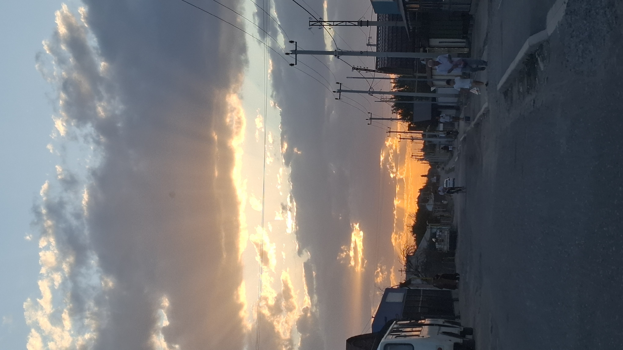 The image depicts a street scene in the city of Shubarssu, Kazakhstan, during sunset. The sky is filled with dramatic clouds, illuminated by the warm orange and yellow hues of the setting sun. Rays of sunlight break through the clouds, casting a soft glow over the scene.
On the left side of the street, a white bus is parked. Further down the street, houses and buildings with flat roofs line both sides. Utility poles with electrical wires are prominent along the right side of the road.
Several people are visible on the street. On the right sidewalk, an elderly man and a young boy are walking, the man carrying a plastic bag. Further up the street, a person is riding a bicycle. Other individuals are scattered along the road and sidewalks, some appearing to be walking. The road surface is paved with asphalt. The overall impression is that of a quiet, residential street in a small town or a suburban area.