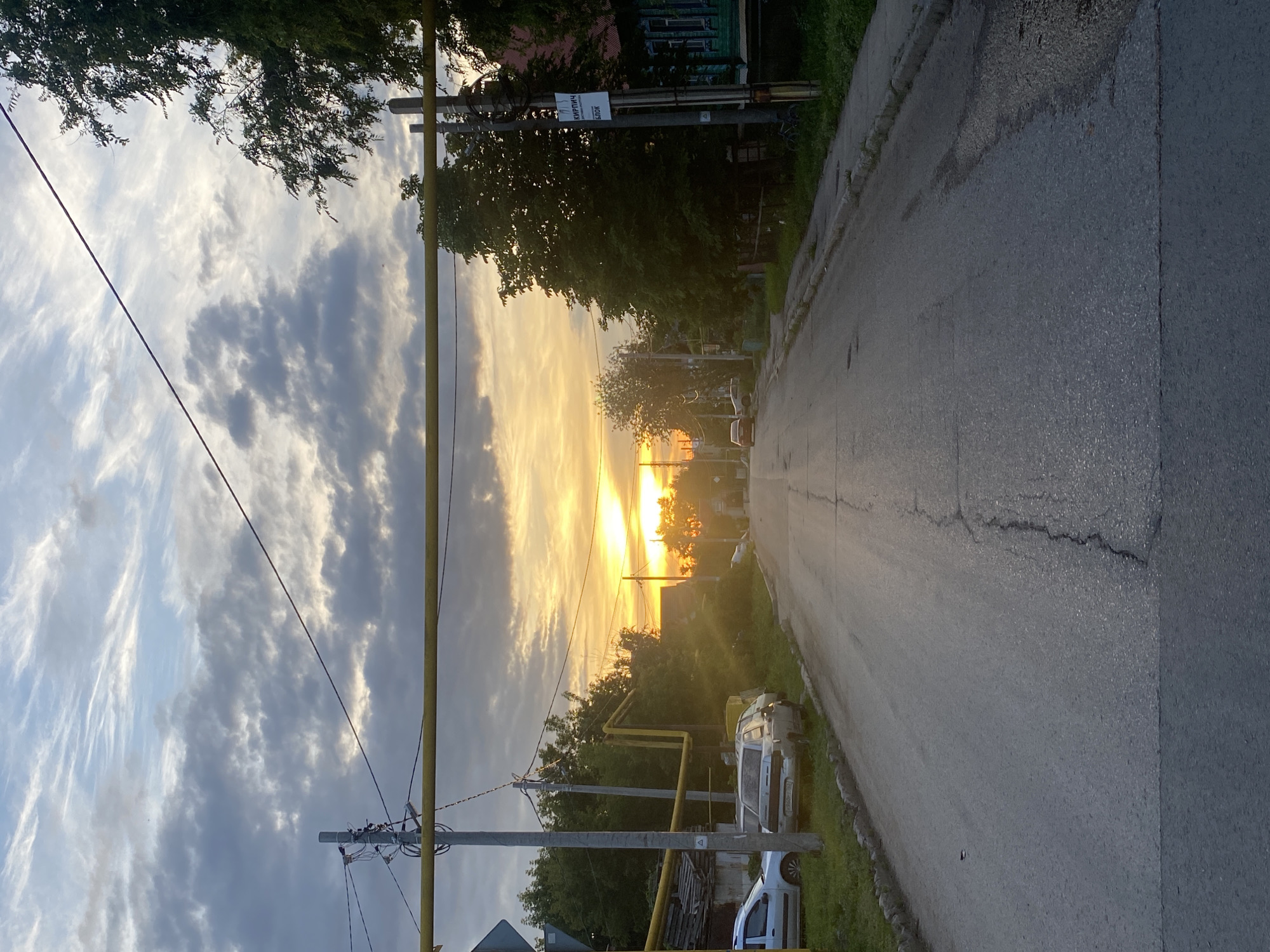The image depicts a rural street scene at sunset in Novokuybyshevsk, Russia. The sun is setting on the horizon, casting a warm, golden glow across the sky and illuminating the clouds with streaks of orange and yellow. Tall trees line the left side of the road, their leaves silhouetted against the vibrant sky. A power pole with wires runs vertically down the left side of the image, a common sight in rural areas. The road itself is asphalt and appears to be in moderate condition, with some cracks visible.
On the right side of the road, there are houses and other buildings visible through the trees. A few parked vehicles, including a white van and a smaller white car, are visible on the grass verge next to the road. Yellow gas pipes run along the ground, further indicating a residential or semi-industrial setting. The overall mood is peaceful and serene, evoking the tranquility of a rural evening. The time of day is clearly sunset due to the warm lighting and low angle of the sun. The weather appears to be fair, with some dramatic cloud formations adding to the visual appeal of the sunset. There is some visible text on a sign attached to the power pole, which appears to be an advertisement, but the text is too small and blurry to be deciphered.