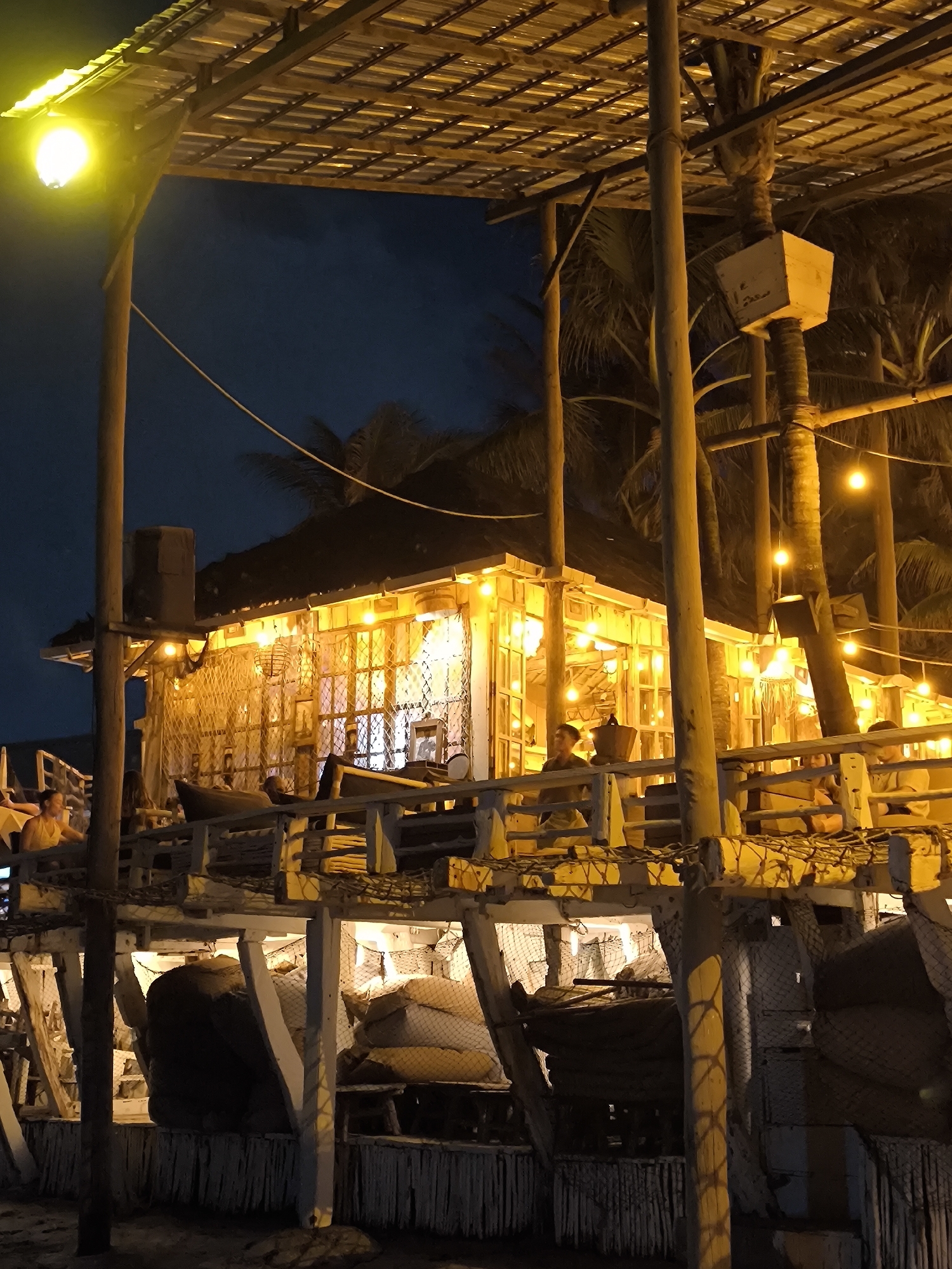 The image depicts a beachfront establishment at night in Canggu, Indonesia. The primary structure appears to be a multi-tiered, elevated wooden building illuminated with warm, yellow lights. People are visible on the upper levels of the building, suggesting it is a restaurant or bar. The lower levels seem to be used for storage, possibly related to fishing, given the presence of nets and large sacks. Palm trees are visible in the background, adding to the tropical setting. The lighting and the overall ambiance create a warm, inviting atmosphere.