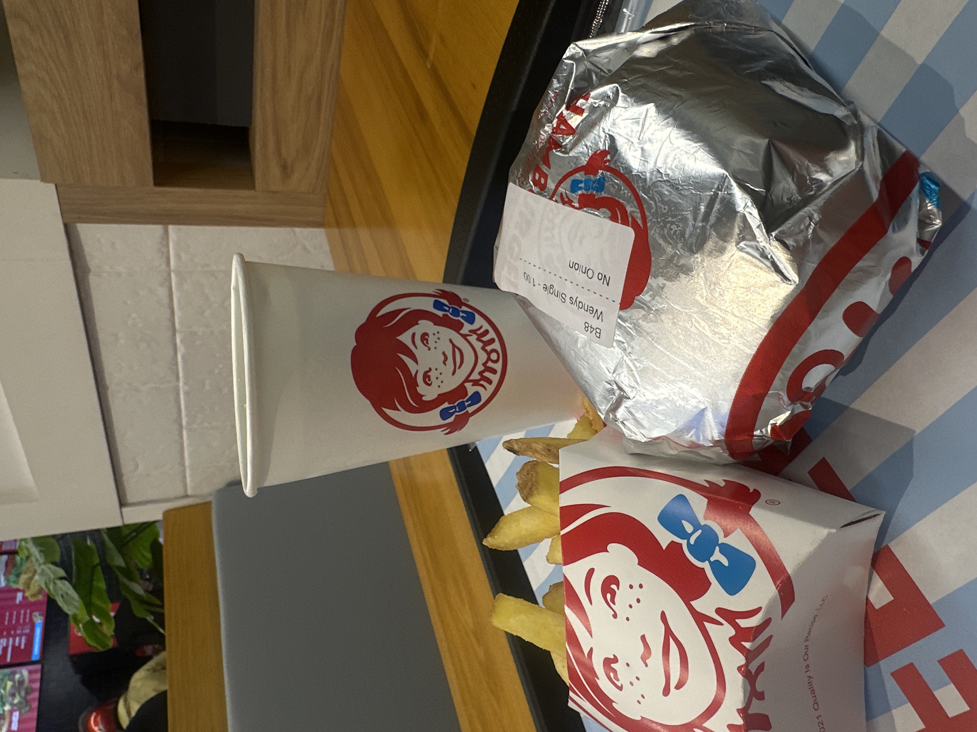 The image shows a Wendy's meal on a black tray. The meal consists of a burger wrapped in foil, a carton of fries, and a drink in a Wendy's cup. The burger wrapper has a label that reads "Wendy's Single - 1.00 No Onion". The meal is placed on a blue and white striped surface. The background includes a wooden panel and a glimpse of indoor plants. The location, Yerevan, Armenia, is not evident from the provided image.