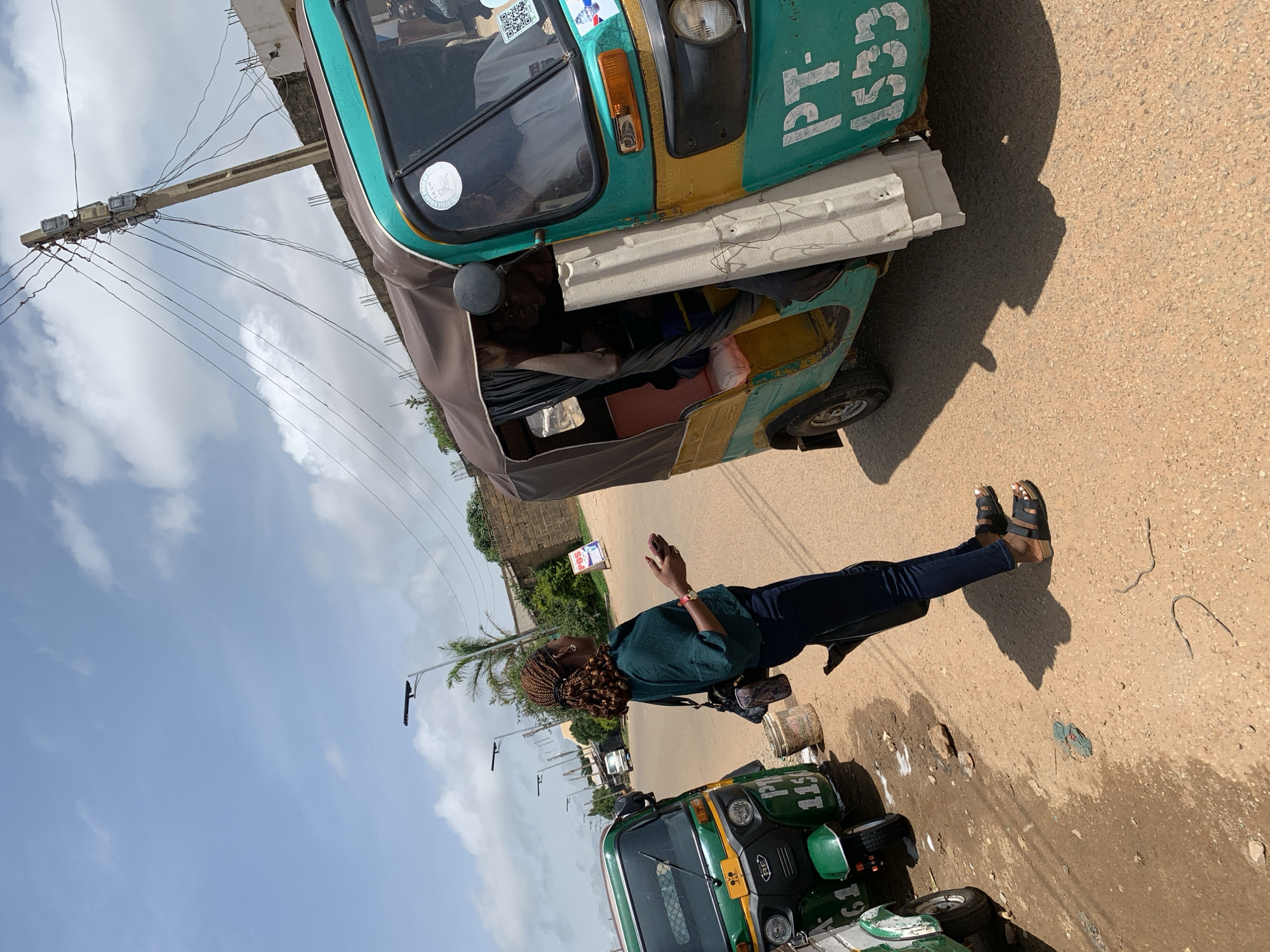 This media file depicts an outdoor scene in Jos, Nigeria, on a bright, sunny day with scattered clouds. The primary focus is a woman standing next to a green and yellow auto-rickshaw, also known as a 'Keke'. She is wearing a dark green shirt, blue jeans, and dark sandals. Her hair is styled in braids, and she is holding a pink mobile phone in her right hand. The woman appears to be looking at her phone.
The auto-rickshaw has the text "PT-1553" written on its side in white lettering. Another, similar auto-rickshaw is visible in the background, partially obscured by the first one. The setting appears to be a street, with a road surface composed of asphalt and gravel. A concrete utility pole with multiple electrical wires is prominent in the background, suggesting an urban or semi-urban environment. Buildings with brick walls are also visible further in the background, along with some greenery and palm trees. The overall impression is a snapshot of daily life on a street in Nigeria, possibly a commercial or transportation hub, given the presence of the auto-rickshaws which are a common mode of transport in the region. The lighting and shadows suggest it is daytime, likely mid-morning or afternoon.