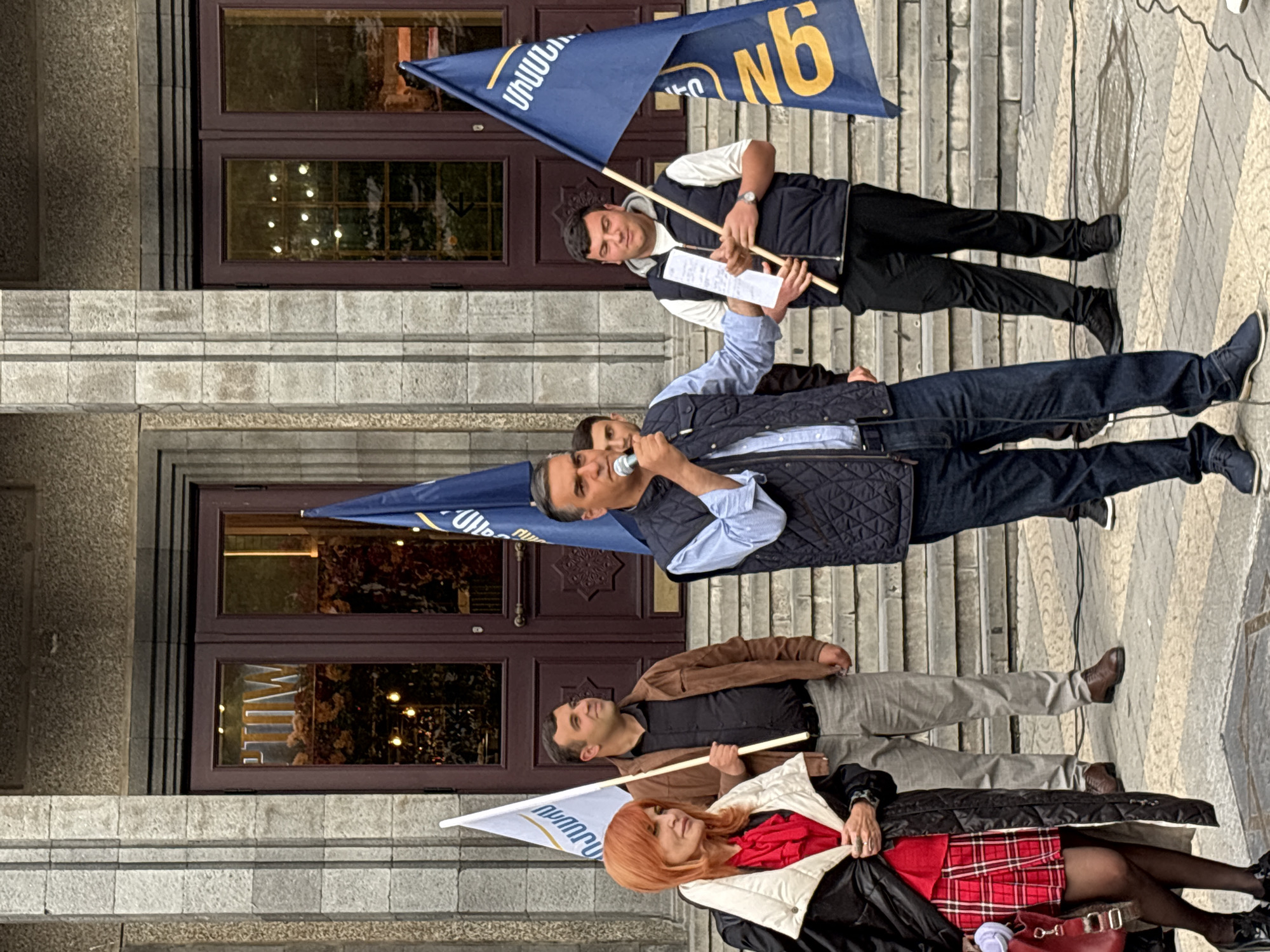 A group of people stand on the steps outside a building. The location is in Yerevan, Armenia, as implied by the flags bearing Armenian script.
The main subject is a man speaking into a microphone. He wears a light blue shirt and a dark quilted vest, and dark trousers. Behind him, a man holds a dark blue flag with a gold-colored "N6" on it. The man with the flag wears a white-sleeved shirt under a dark vest, with dark trousers.
Below the speaker, a man in a brown jacket and grey trousers holds a white flag with Armenian text. Beside him is a woman with reddish-orange hair. She wears a black jacket over a red top and a red plaid skirt.
The steps are grey stone. The building has dark-framed glass doors and windows, with visible reflections of lights and possible interior decor.