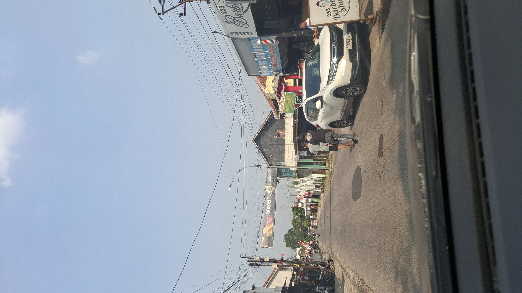The image depicts a street scene in Sukasari, Indonesia, on a clear day with a bright blue sky and scattered clouds. The perspective is from inside a vehicle, looking forward. The road is paved and appears to be a moderately trafficked local street. On the left side of the road, there are buildings and parked motorcycles. Flags and banners are visible along the roadside, suggesting a local event or celebration.
In the center of the road, two men are walking away from the camera. A white Honda CR-V is parked on the right side of the road, partially obscuring the storefronts behind it. The storefronts on the right appear to be commercial establishments, one of which is a barbershop identified by a sign that reads "233 Barber Shop." A man is standing near the entrance of the barbershop. Another banner is stretched across the street in the distance, displaying text and an image of bananas, likely an advertisement. Overhead, electrical wires are strung across the street, supported by utility poles. The overall scene suggests a typical urban or suburban street in Indonesia, with commercial activity and pedestrian and vehicular traffic.