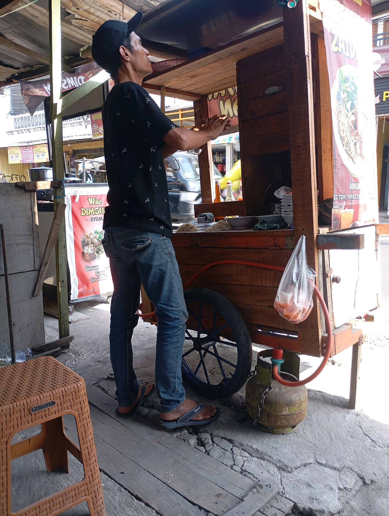 This is a full-shot, eye-level, outdoor, daytime, eye-level photograph of a man standing next to a food cart at a market stall in Babelan, Indonesia. The man, wearing a black baseball cap, black t-shirt with a white patterned print, blue jeans, and brown flip-flops, is looking upwards and to the right, with his left arm raised as if to gesture or adjust something above him. He appears to be a vendor at the food cart, which is made of wood and has a metal wheel on the left side. A gas cylinder is attached to the cart. To the left of the cart is a brown plastic stool. Behind the cart, there are other market stalls, some with banners and goods displayed. The ground is a mixture of concrete and wooden planks. The lighting suggests a sunny day.
