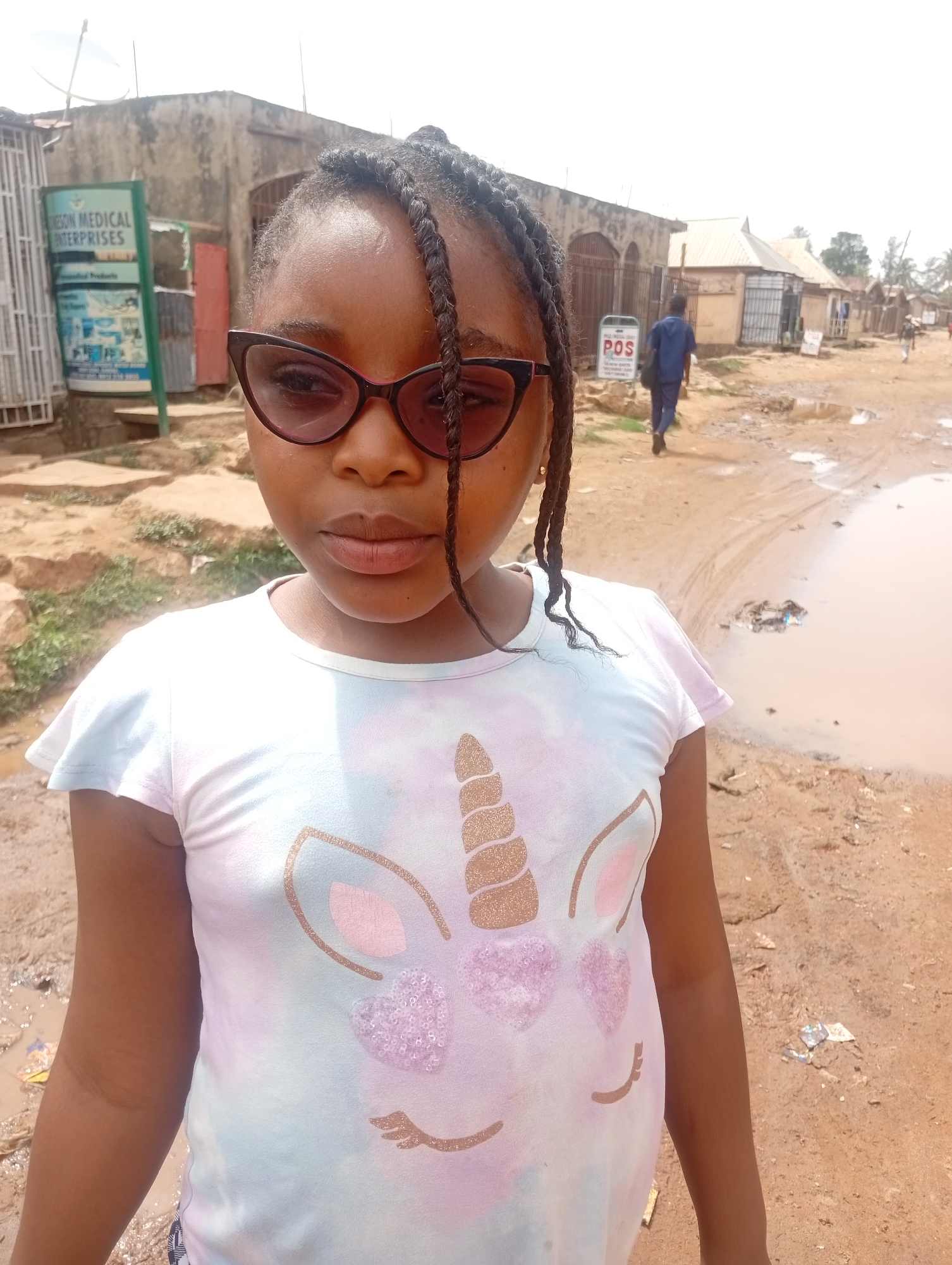 A young girl with braided hair and sunglasses stands in a residential area in Sabon Tasha, Nigeria. She is wearing a t-shirt with a unicorn design. Behind her, a dirt road is partially flooded with muddy water. Buildings and a sign reading "KESON MEDICAL ENTERPRISES" are visible in the background. A man in a blue shirt is walking away in the distance.