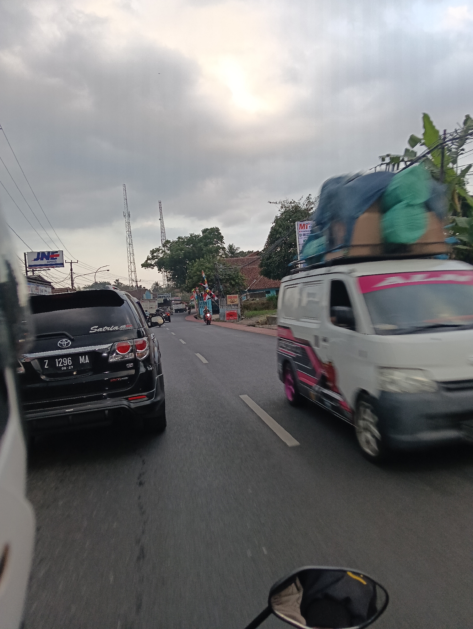 The image depicts a street scene in Linggajaya, Indonesia, on a cloudy day. The perspective is from a moving vehicle, likely a motorcycle, as indicated by the motorcycle's rearview mirror in the foreground, reflecting a person wearing dark clothing. The road is paved and has white lane markings.
In the foreground, to the left, is the rear of a black SUV. Its license plate reads "Z 1296 MA". The SUV has a "Satria" sticker on its rear. To the right of the SUV, a white van is moving in the same direction. The van has colorful decals on its side and is carrying a load of green and brown items secured on its roof with blue tarpaulin.
Further down the road, several other vehicles are visible, including trucks and motorcycles. On the left side of the road, a sign for "JNE EXPRESS" is mounted on a pole. Two tall communication towers are visible in the distance against the cloudy sky. On the right side of the road, there are trees, houses with red-tiled roofs, and some flags. The overall impression is a busy road in a semi-urban or rural Indonesian setting.