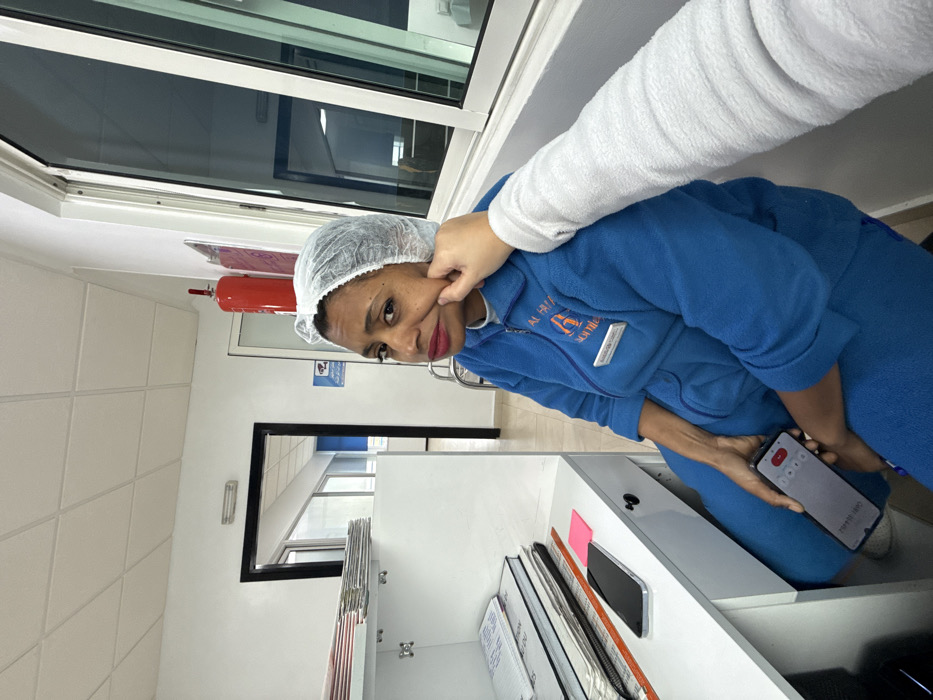 An indoor scene depicts an adult female seated in what appears to be a workplace environment. The woman, characterized by medium-dark skin, dark hair covered by a white disposable hairnet, is wearing a blue zippered uniform or tracksuit. A name badge, displaying "AL HU..." and "A somile," is clipped to her chest. Her face, adorned with red lipstick, is turned towards the viewer, showing an exaggerated expression with pursed lips, while an arm covered in a white textured sleeve, extending from the right foreground, gently pinches her left cheek. She holds a black smartphone in her left hand, which displays an audio recording interface.
The surrounding environment consists of white walls and a white grid-patterned ceiling. A red fire extinguisher is mounted on the wall, adjacent to a blue sign depicting an eye symbol (indicating CCTV surveillance). Large windows occupy the right side of the frame. To the woman's left, a white desk unit holds stacked papers and notebooks on its shelves, with a second black smartphone resting on a lower surface. The specific location within the city of Meknes, Morocco, is not visually identifiable from the image.