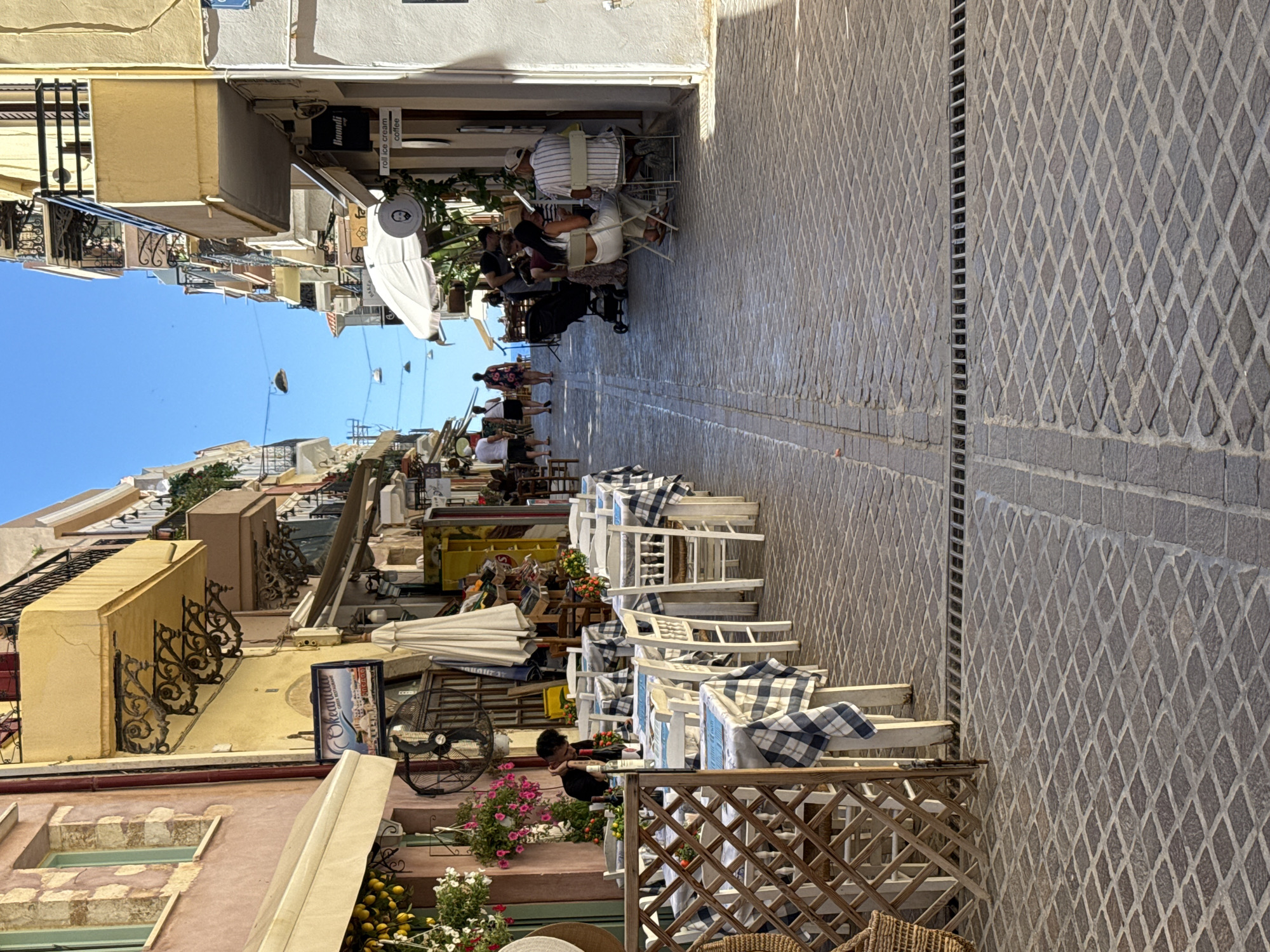 The image captures a narrow, cobblestone street in Chania, Greece, lined with buildings and outdoor dining establishments. On the left, a pink building with arched windows and a cream-colored awning is visible. A sign for "Okeanida Greek Restaurant" hangs on a yellow building further down the street. Several tables with blue and white checkered tablecloths are set up on the right side of the street, occupied by diners. A person wearing a black shirt sits alone at one of the tables. Further down the street, more tables and people are seated, some under a large white umbrella. The street is paved with diamond-shaped cobblestones, and a metal grate runs across the middle. The sky is a clear, bright blue. The overall scene depicts a vibrant, Mediterranean atmosphere, typical of the old town in Chania.