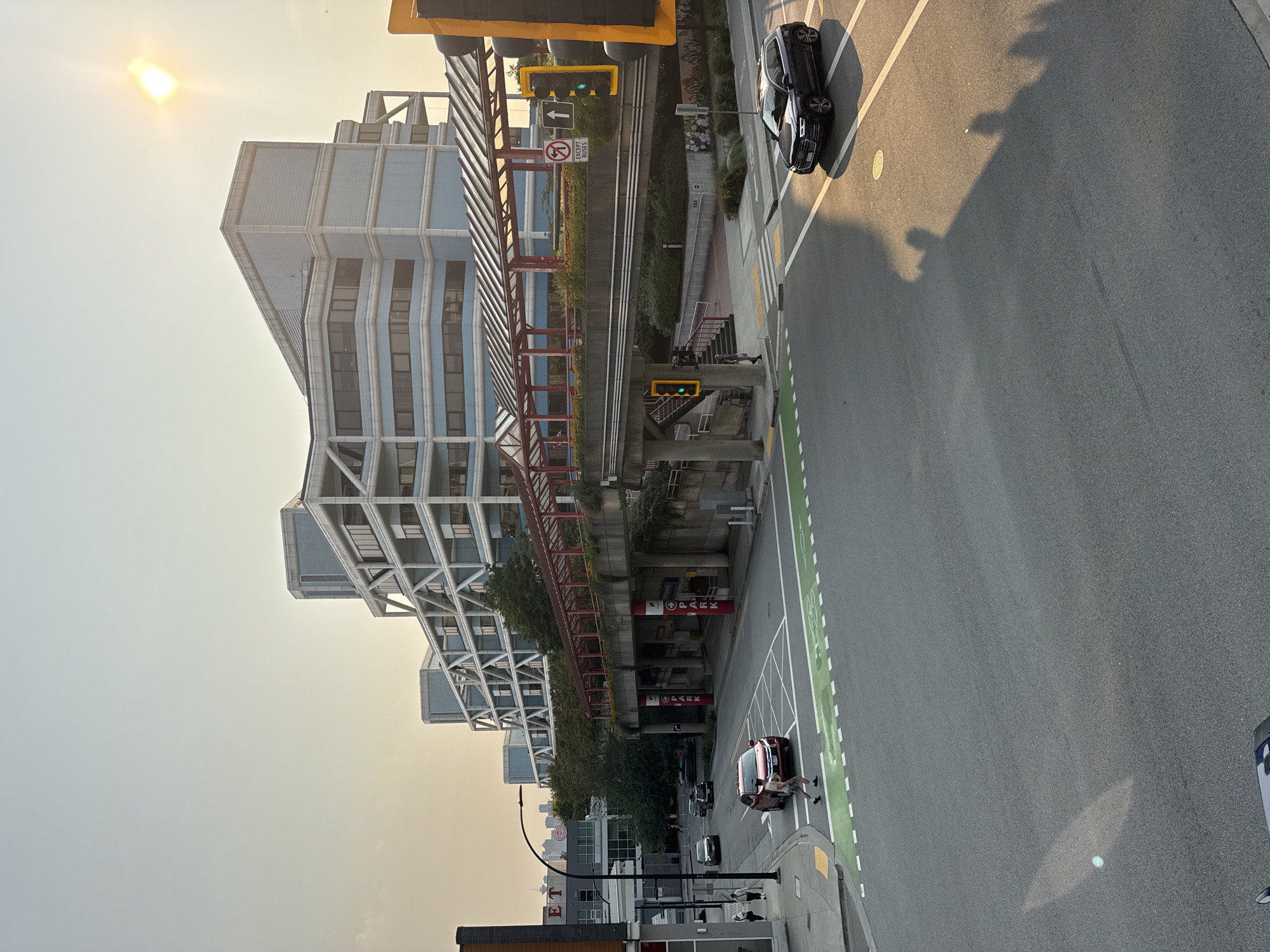 The image captures a street scene in North Vancouver, Canada, likely during late afternoon or early evening due to the low angle of the sun casting long shadows. In the background, a modern, multi-story building with a distinctive white, angular exoskeleton structure dominates the skyline. The building is clad in pale blue and grey materials, with numerous windows.
In the midground, a covered pedestrian overpass with a red frame and a translucent roof spans across the street. The overpass is supported by concrete pillars, and visible signage on the pillars reads "PARK" in red lettering. Lush green foliage surrounds the base of the overpass and lines its edges.
The foreground shows a city street with asphalt pavement. A pedestrian crossing with green markings is visible. A red sedan is stopped at the crossing, and a person in shorts and a t-shirt is walking across the street on the pedestrian crossing. A dark-colored SUV is also present on the road, with its shadow cast onto the asphalt. Traffic lights with illuminated green signals are visible at the intersection. The sky is a hazy, light orange-yellow, suggesting a possibly smoggy or sunset atmosphere. The overall impression is of an urban environment with modern architecture, pedestrian infrastructure, and active vehicular and foot traffic.