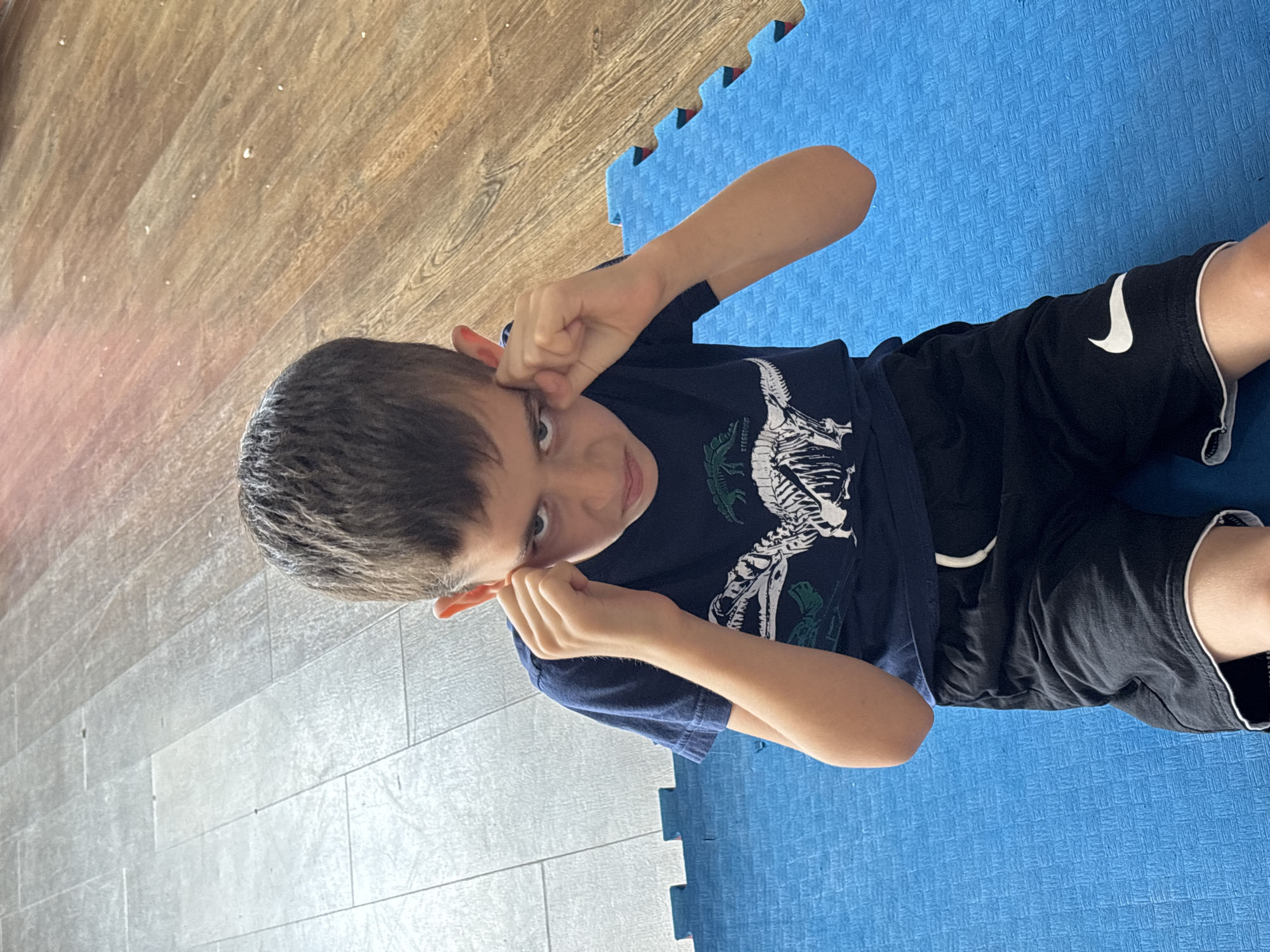 A young boy, appearing to be of Caucasian ethnicity, sits on a blue puzzle mat in a room with wooden flooring and a tiled floor. He is wearing a dark blue t-shirt with a white dinosaur skeleton graphic and black shorts with a white Nike logo. His hands are raised to his temples, and he has a determined or angry expression on his face. The lighting suggests an indoor setting, possibly during the daytime. The specific location within Chaikovsky, Russia, cannot be determined from the image alone.