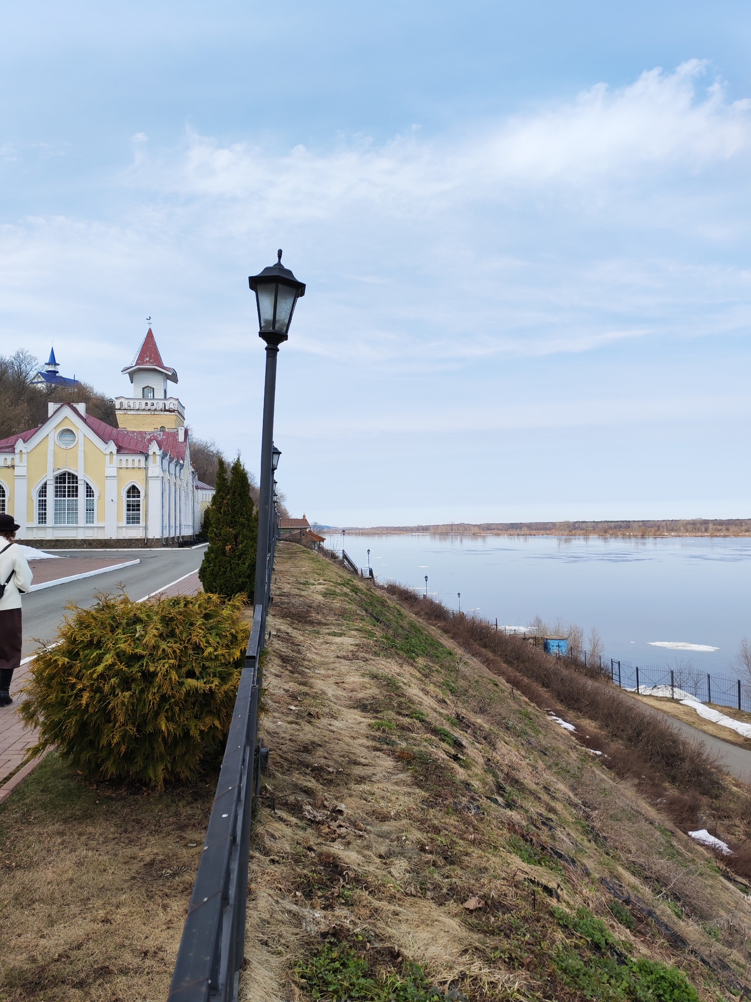 The image shows a view in Sarapul, Russia. A yellow and white building with red and blue architectural features stands on a hill. To the right, a black metal fence runs along a grassy slope leading down to a body of water. A black lamppost stands near the fence. A paved path runs alongside the building. A person wearing a white coat and brown pants is walking near the building. The sky is light blue with scattered clouds. The water shows signs of ice.
