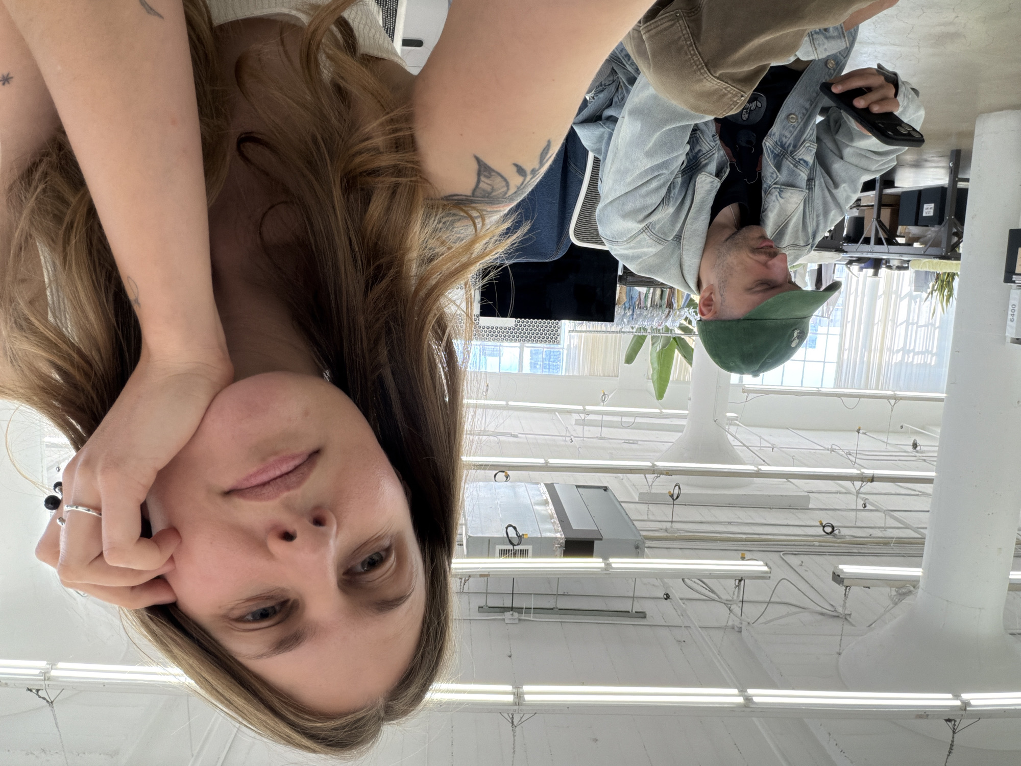 The image shows two people in a bright, white, industrial-looking space. A woman with long brown hair is in the foreground, her face close to the camera and resting on her hand. She has fair skin and blue eyes. Behind her, a man is visible, wearing a green baseball cap and a denim jacket. He is holding a phone in his hand. The background reveals a high ceiling with numerous fluorescent lights and some ductwork. Through a window in the background, it's possible to discern elements of a cityscape, which suggests an urban setting. Given the architectural details, it could potentially be located in an industrial area of Los Angeles, United States.