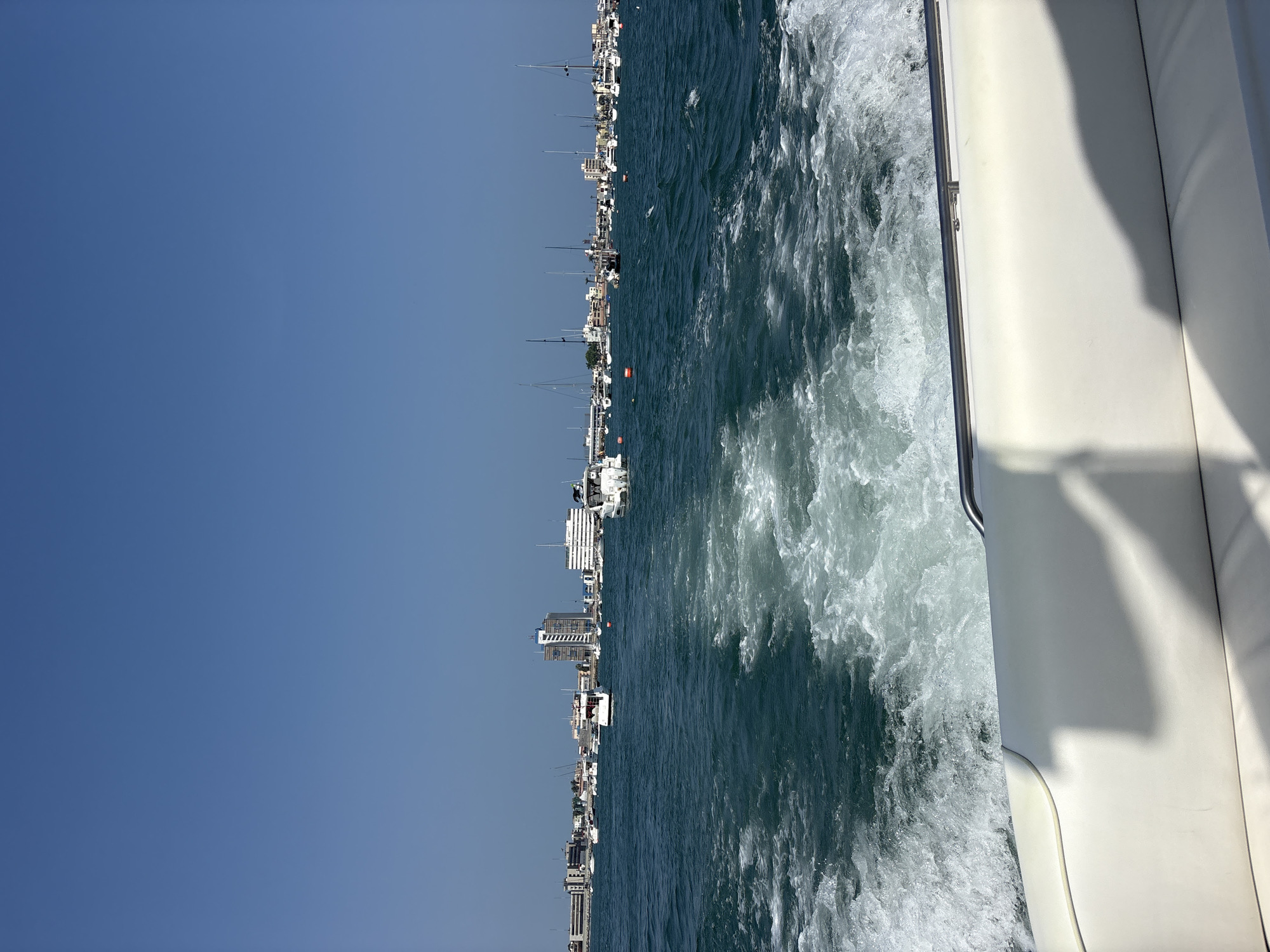 The image captures a scene from the perspective of someone on a boat, likely moving through the water. The wake of the boat is visible on the right side of the frame, with turbulent water and foam. The water is a deep blue-green color. In the distance, along the horizon, lies the cityscape of La Punta, Peru, with visible buildings, including high-rise structures, and several boats and sailboats anchored near the shore. The sky is a clear, bright blue. On the right, a portion of the boat's white structure is visible.