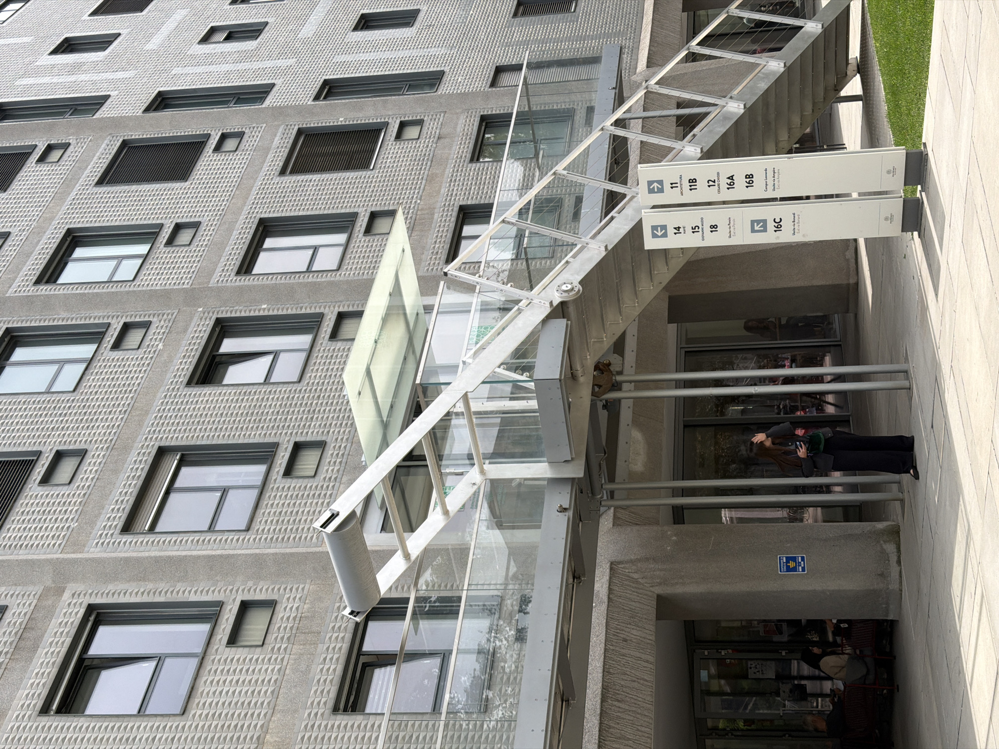 Here is a detailed description of the image:
The image captures the exterior of a modern building, likely in Milan, Italy, as indicated by the Italian language signage. The building has a distinctive facade with a patterned surface and recessed windows. An external steel staircase and a glass-enclosed elevator are attached to the building's exterior.
Signage is prominently displayed, with directional arrows and names like "ARCHITETTURA," "CESARE CHIODI," and "Campus Lesnardo," suggesting the building is part of a university or campus complex. Additional text indicates directions to "Uscita via Panzio," "Uscita via Ampère," and "Uscita via Bonardi," which suggests exit routes to nearby streets.
A woman stands near the entrance of the building. She wears dark clothing, and is looking at a cell phone. There is a second person near her, also looking at a cell phone.
The environment includes stone or concrete paving and some greenery near the top of the image, indicating landscaping. The scene is well-lit, implying it is daytime. The overall impression is of a contemporary academic or institutional building in an urban setting.