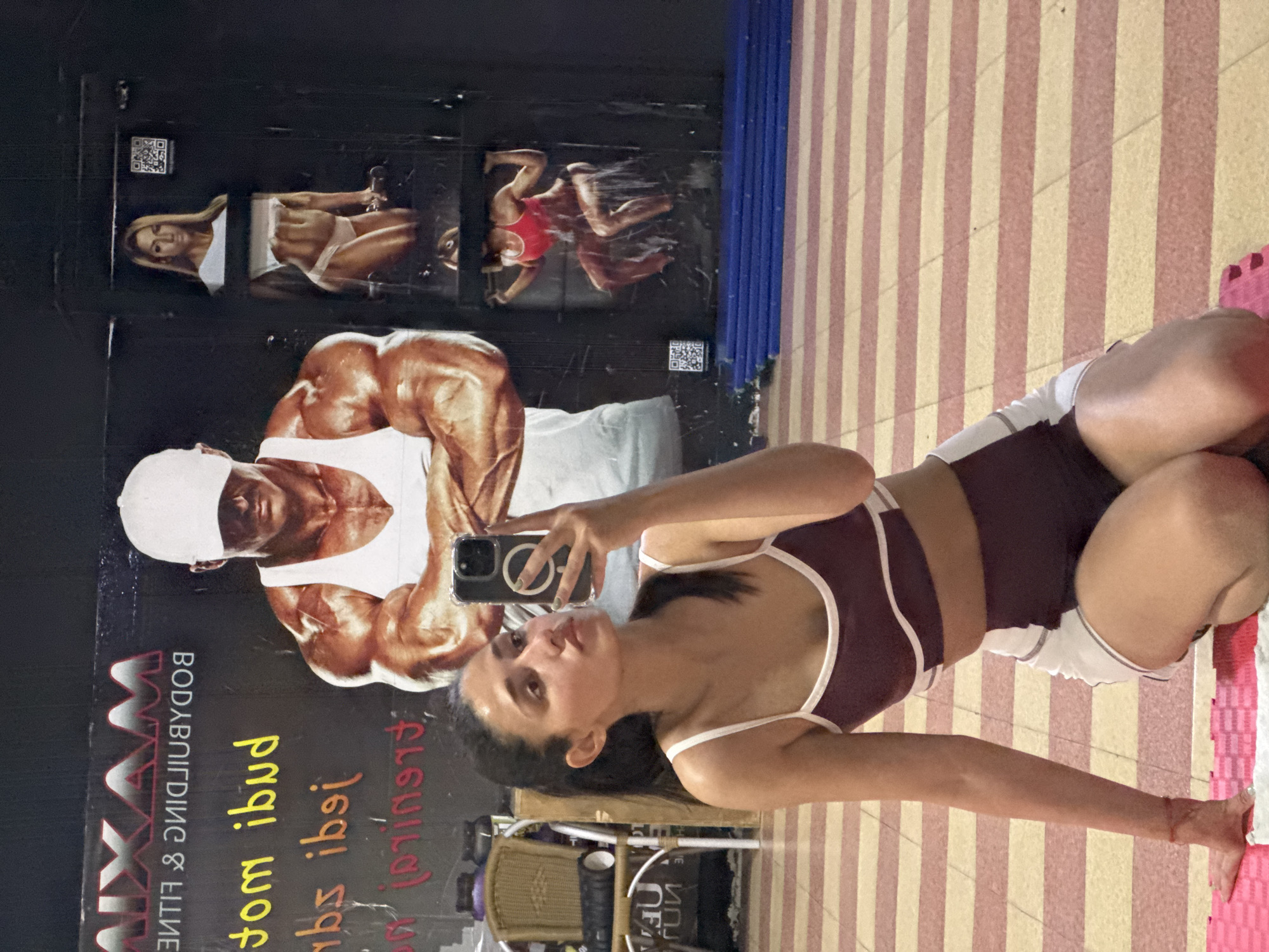 A woman is taking a selfie in a gym in the city of Bar, Montenegro. The gym has a floor covered in pink and cream striped mats. There are posters on the walls of bodybuilders. The woman is wearing a two-piece athletic outfit.