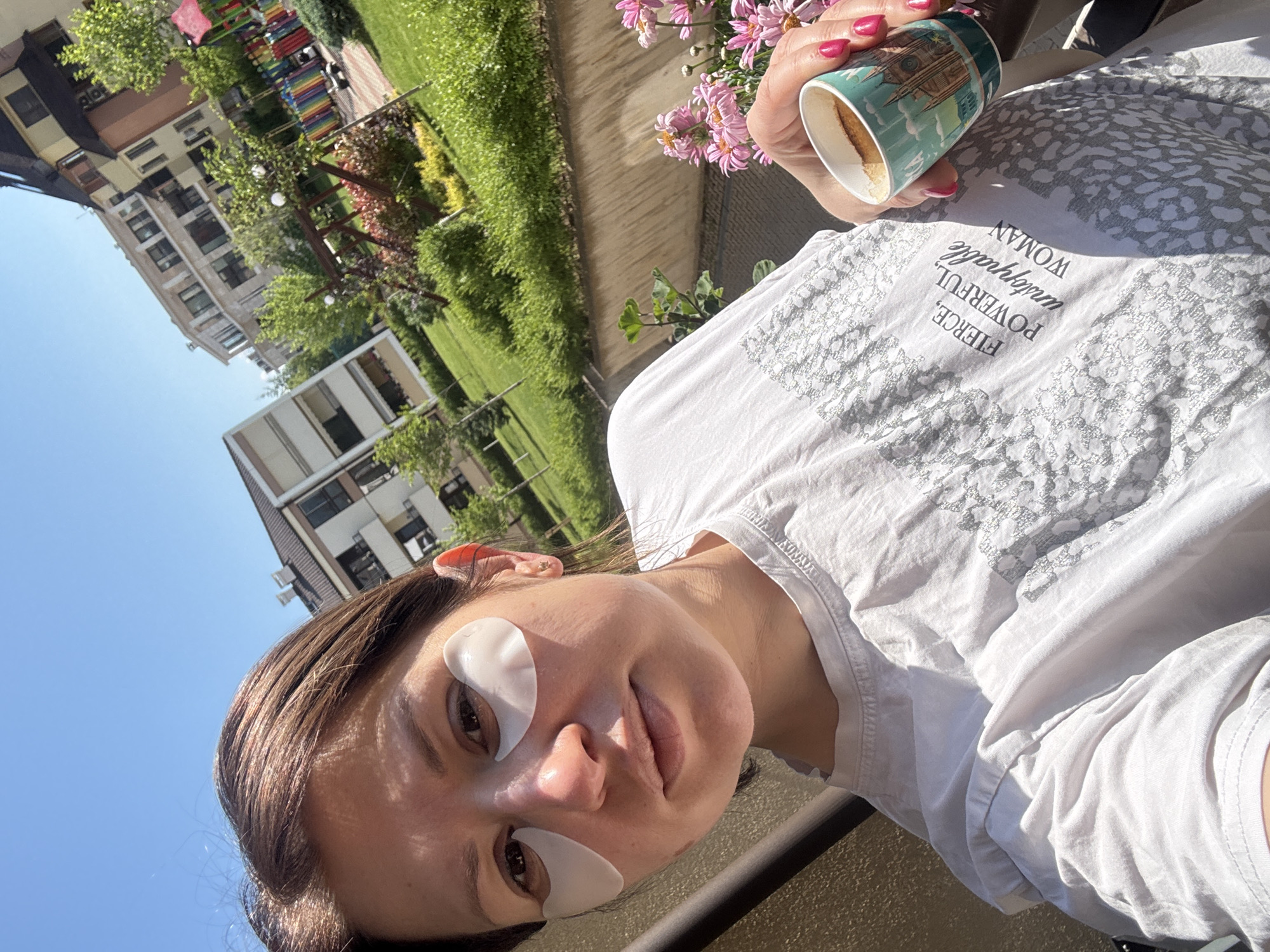 A woman with eye patches is pictured holding a coffee mug in what appears to be an outdoor setting. The woman is wearing a white t-shirt with text. The backdrop features greenery, buildings, and a view of the sky. Considering the architectural style and the general surroundings, the location may be Sofia, Bulgaria. There are pink flowers visible. A mug with coffee is held by a hand with pink nail polish.